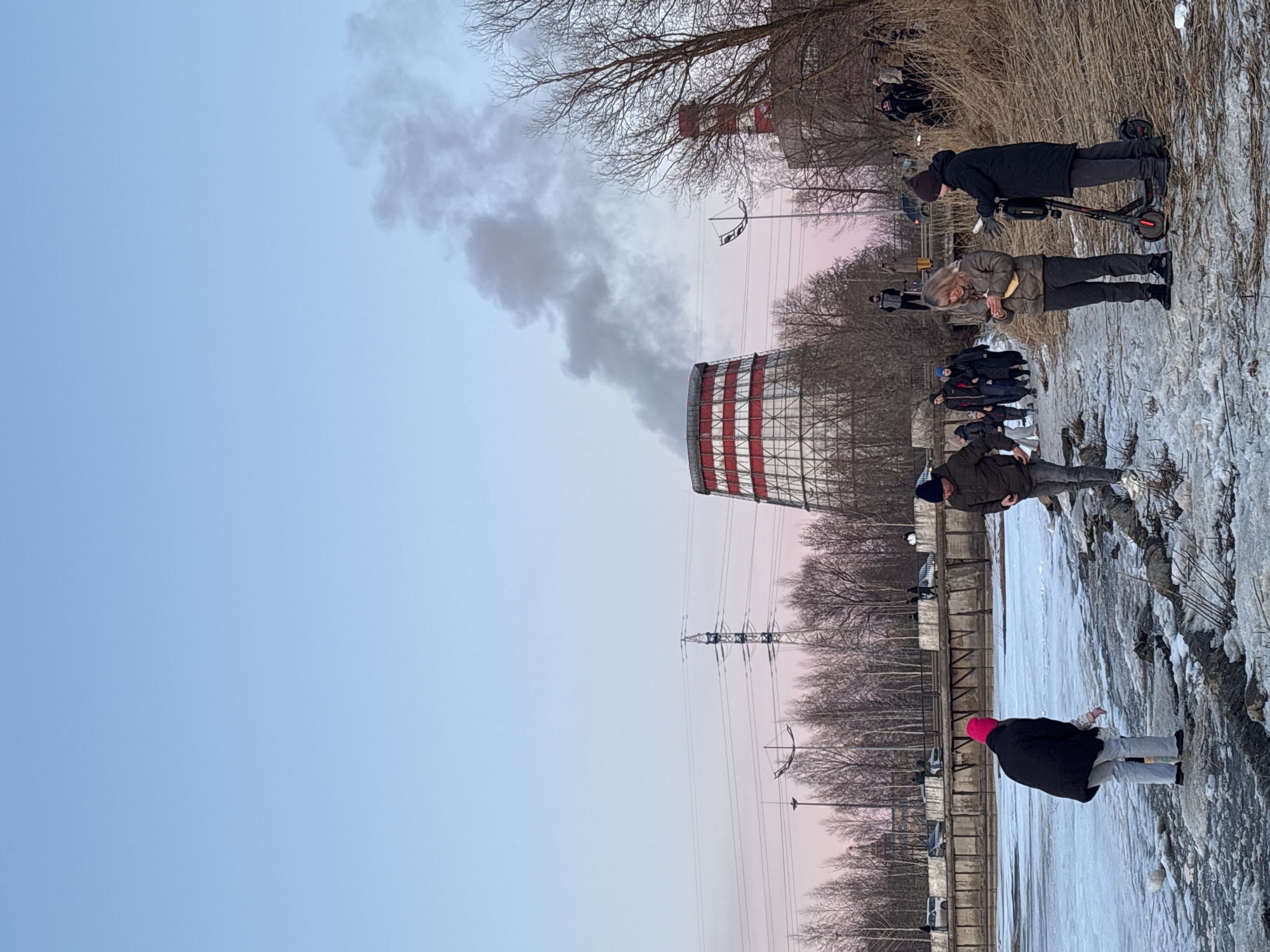 The image shows a group of people in a wintry outdoor setting in Izhevsk, Russia. A red and white striped cooling tower is visible in the background with smoke rising from it. A smaller red and white structure is visible in the distance behind a line of bare winter trees. Overhead power lines are also visible in the background. The foreground consists of a frozen or icy surface. Several individuals are walking or standing on this surface. One person is riding a scooter. Another person wears a pink hat. The sky is pale blue and pink.