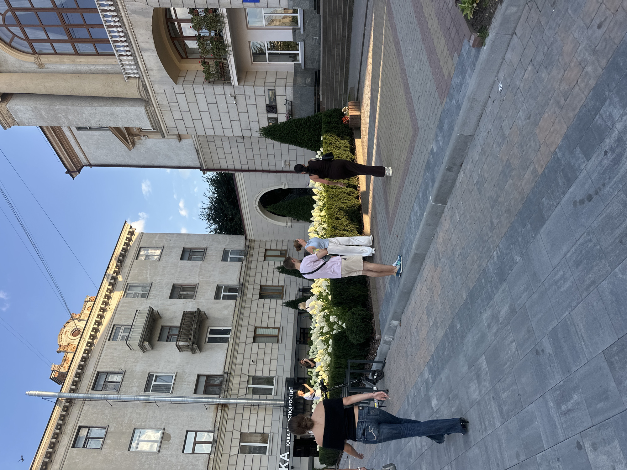 This image depicts a street scene in the city of Ternopil, Ukraine, specifically on Ternopil's main street, Soborna Street, in front of the Ternopil Regional State Administration building. The photograph captures several individuals and architectural elements characteristic of an urban environment.
In the foreground, a woman with dark hair is walking away from the camera, wearing a black off-the-shoulder top and blue jeans. To her left, a parked electric scooter is visible. Further into the scene, a group of people stand near a large flowerbed filled with white hydrangeas. One person, a man wearing a pink t-shirt and light-colored shorts, is facing the flowerbed. To his right, two other individuals are also looking towards the flowers.
The background features two prominent buildings. The building on the left is a multi-story apartment building with numerous windows and balconies, exhibiting a somewhat weathered facade. To the right is a grander, lighter-colored building with architectural columns and a large arched window. Both buildings are indicative of Soviet-era architecture. The sky above is clear and blue, with a few wispy clouds.
The ground is paved with interlocking concrete blocks in various shades of gray, brown, and red, forming a decorative pattern in some areas. A curb separates the pedestrian walkway from the street. The overall atmosphere suggests a pleasant day in a public urban space.