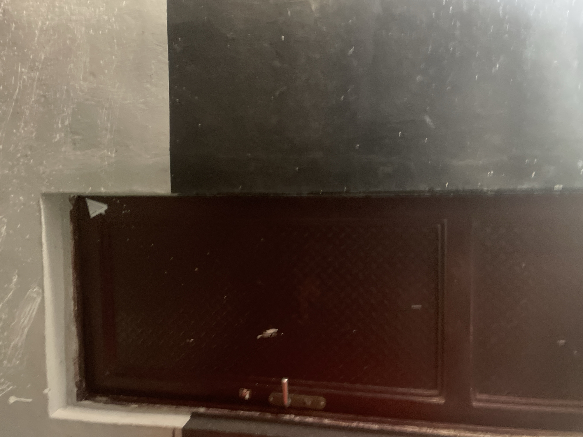 The image shows a close-up view of a dark brown door with a metal handle. The door appears to be made of metal and has a diamond-shaped pattern embossed on its surface. The wall to the right of the door is painted in two sections: the upper part is a light gray with visible scratches and smudges, while the lower part is a dark gray or black. The door is situated within a doorway that has a white frame. The overall environment suggests an interior, possibly a hallway or an entrance to a building, in Jauro Yinu, Nigeria.