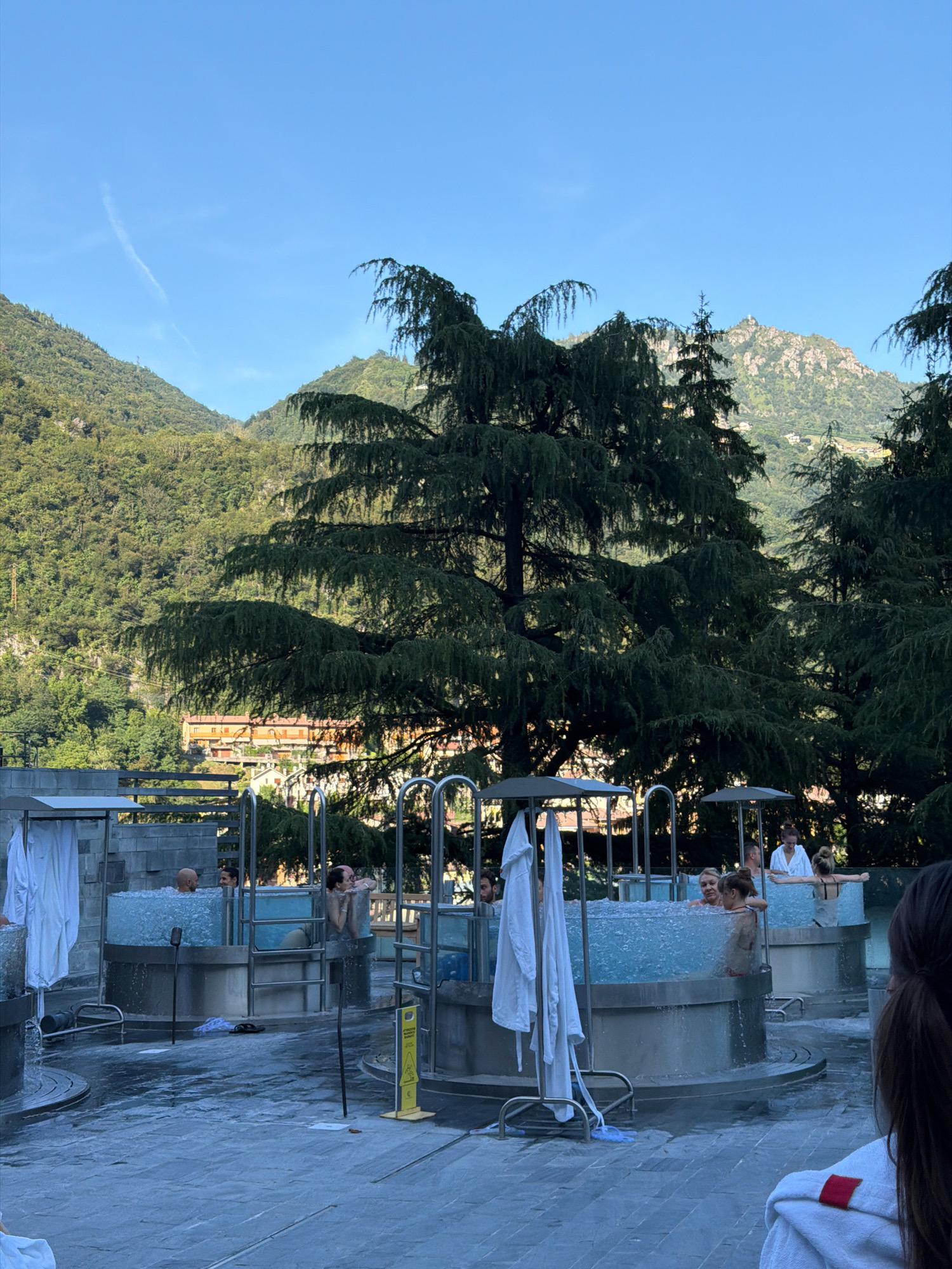 The media file depicts an outdoor thermal spa with several circular, clear-walled pools filled with bubbling water. Several people are seen relaxing in the pools, some conversing while others soak. The setting is mountainous, with lush green hills and trees forming a backdrop. A building with orange facades is visible further up the hillside, suggesting a resort or hotel. Large evergreen trees, possibly cedars, are prominent in the foreground, their branches creating a natural canopy. The pools are surrounded by a paved area, and white robes are hanging on racks near some of the pools, indicating the nature of the establishment. A yellow warning sign with black text and a symbol of a person slipping is visible on the right side, cautioning about wet floors. The sky is a clear, bright blue, and the sunlight suggests it is daytime, likely late afternoon or early evening given the long shadows. The overall atmosphere appears tranquil and leisurely.