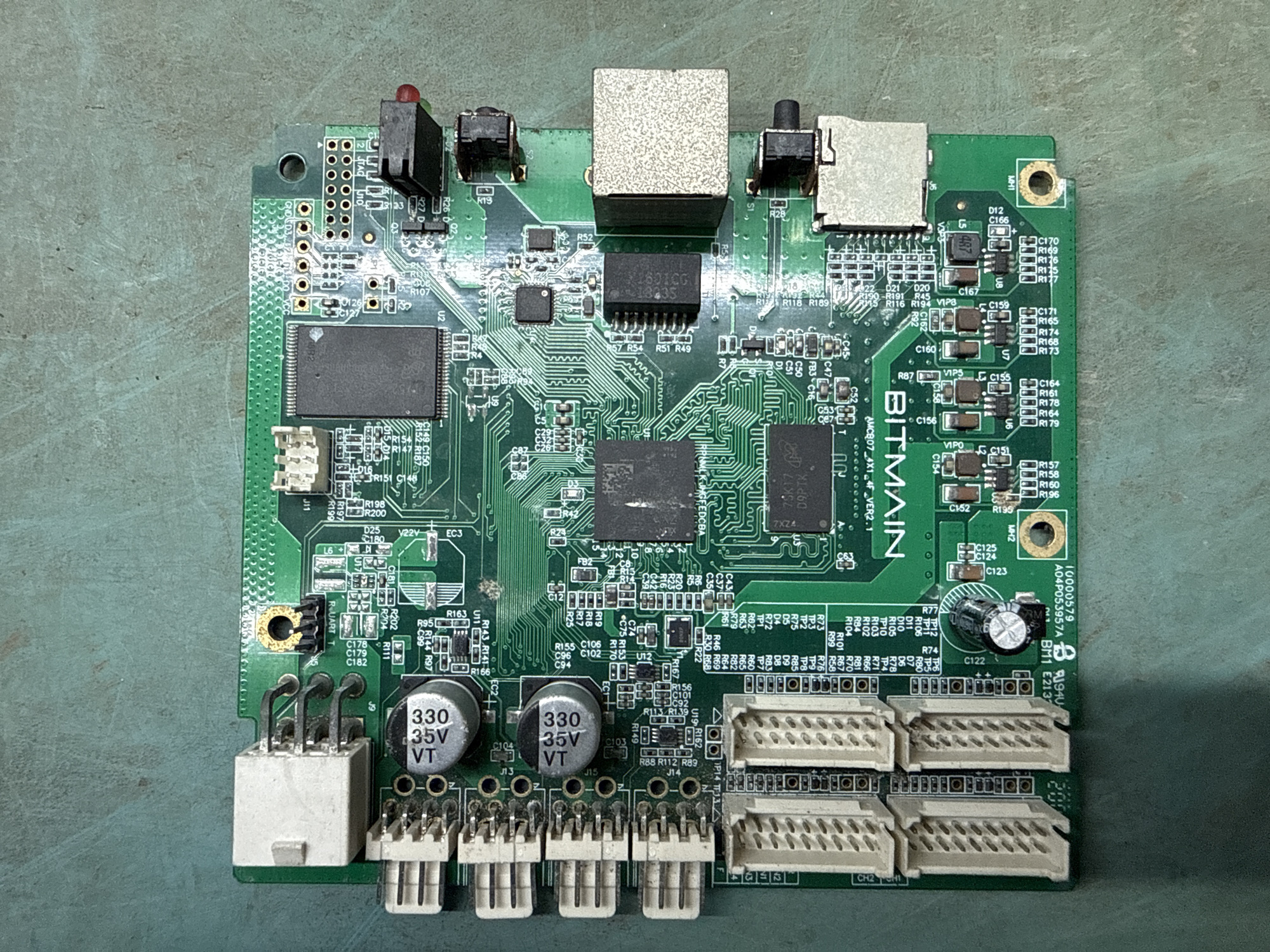 The image displays a green printed circuit board (PCB) with various electronic components. The PCB is laid flat on a textured, greenish-gray surface, suggestive of a workbench or industrial setting. The board is oriented with its longer side running horizontally across the frame.
Prominent components include several white connector blocks with multiple pins, arranged in rows along the left and bottom edges of the PCB. There are also two cylindrical silver capacitors with black tops, each labeled "330 35V VT". A large black integrated circuit (IC) with text that appears to read "1487CC" is centrally located, with another smaller black IC nearby. A prominent white rectangular connector with pins is situated at the top left. There are also several smaller black components resembling surface-mount ICs and resistors.
Towards the right side of the PCB, there are two metallic-looking connectors with square openings, one larger than the other. A small red LED is visible near the top right corner. Text printed on the PCB is visible in several areas, including "BITMAIN" in large white letters near the bottom center, and what appears to be a model or serial number starting with "10000579" near the bottom left. There are also many smaller alphanumeric labels identifying components (e.g., R102, C179, U2, J11).
The overall impression is that of a piece of electronic hardware, possibly a control board or module, photographed for identification or inspection.
This image does not contain any visual information that would allow for the determination of its location in Indiantown, United States of America.