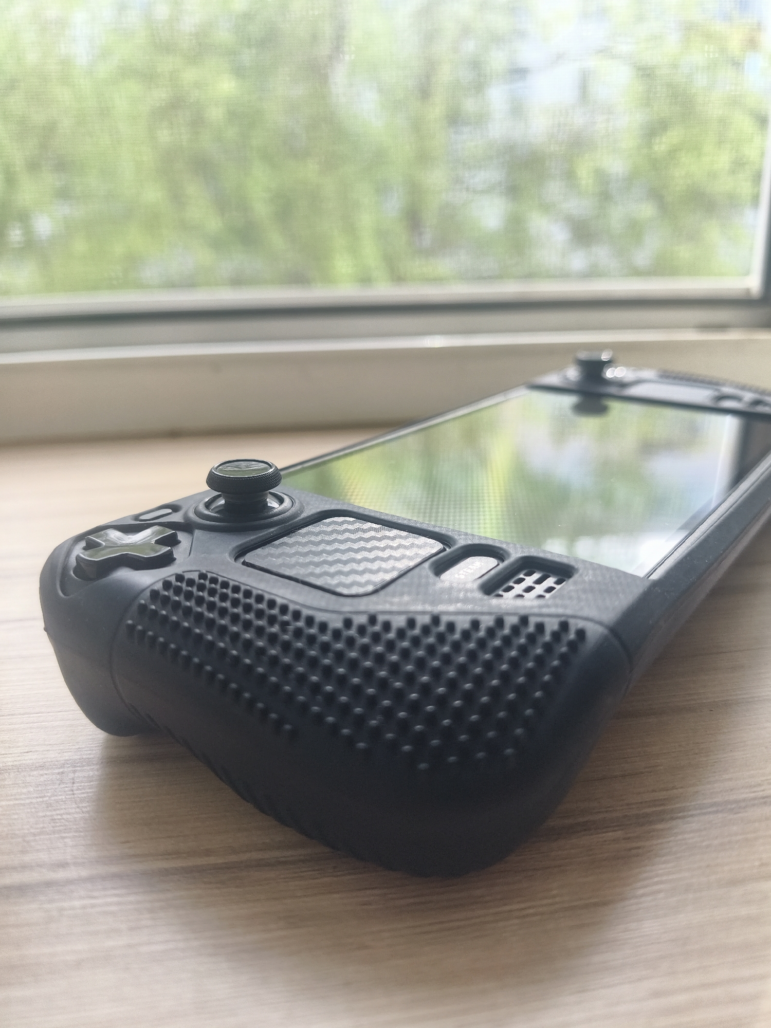 A handheld gaming console sits on a light-colored wooden surface, possibly a table or countertop. The console has a black protective case with a textured grip on the left side and a screen that reflects an outdoor scene with trees visible through a window. A directional pad and analog stick are visible on the left side of the console, and what appears to be the top of another analog stick is visible on the right. The room is illuminated by natural light. The location in the city of Chaikovsky, Russia is not evident.