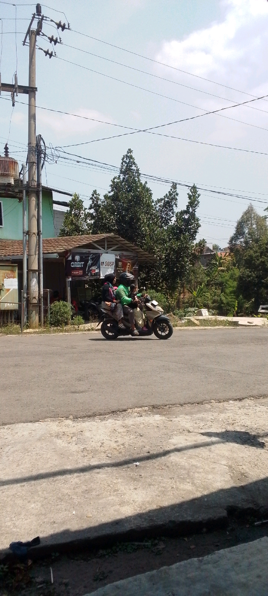In the city of Cilaku, Indonesia, on a clear day, two individuals are riding a motorcycle on an asphalt road. The person in the driver's seat wears a green shirt and a helmet. The passenger, a woman, wears a headscarf and a patterned skirt. They are traveling from left to right across the frame. The background features lush green trees and vegetation, along with a utility pole and power lines. A building with a tiled roof is visible behind the trees. The foreground consists of a concrete sidewalk and some greenery.