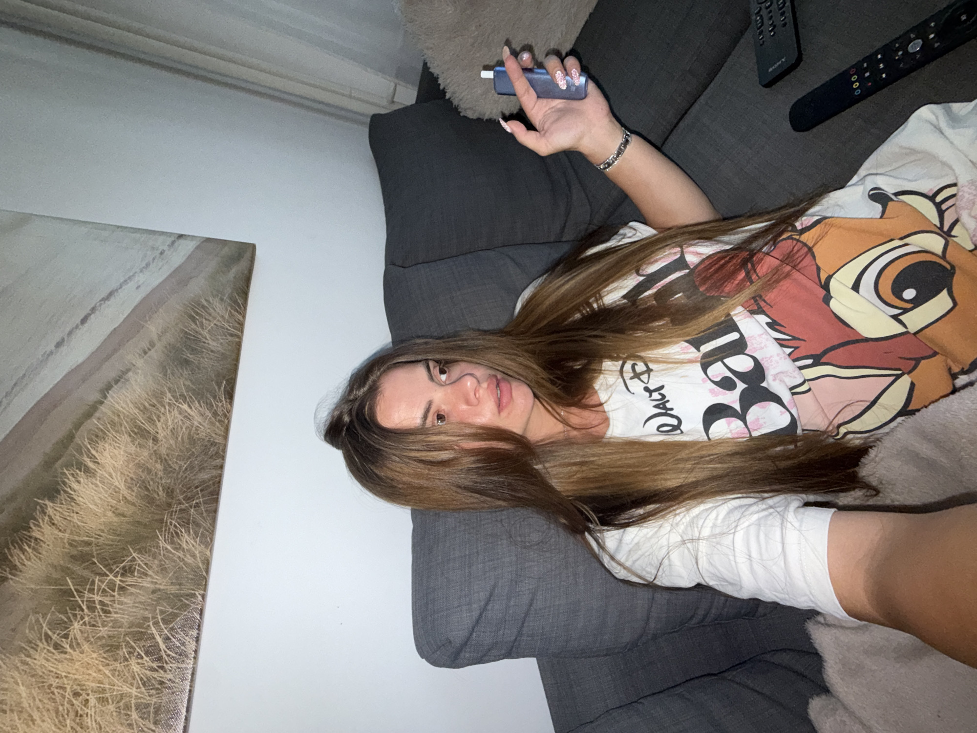 A young woman with long, light brown hair is sitting on a dark gray sofa. She is wearing a white t-shirt with a cartoon print of Bambi. She is holding a small blue electronic device in her right hand. Her left arm is resting on a fluffy beige pillow. There are two remote controls on the sofa next to her. In the background, a large abstract painting with shades of beige and brown hangs on the wall. The room appears to be a living room.