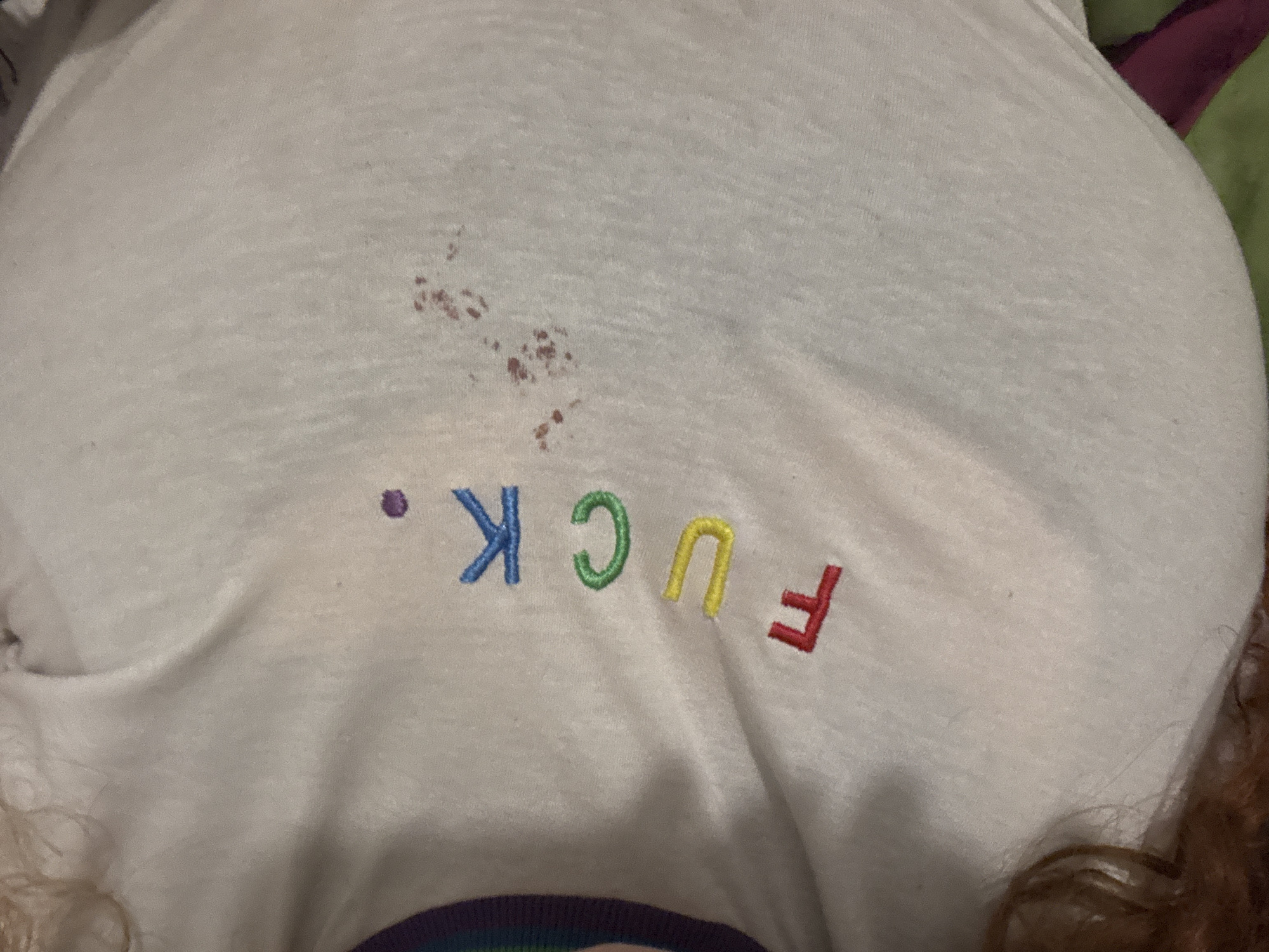A close-up, top-down view captures a white t-shirt being worn, likely by an individual in Buenos Aires, Argentina. The t-shirt fabric appears soft, possibly cotton. A prominent cluster of reddish-brown, irregularly shaped stains is visible on the upper-middle section of the shirt, suggesting a spillage or accident. Below these stains, the word "FUCK." is embroidered in vibrant rainbow colors: the 'F' in red, 'U' in yellow, 'C' in green, 'K' in blue, and the period in purple. Part of a person's brown, curly hair is visible in the bottom right corner of the frame. The background, out of focus, shows hints of other fabrics or clothing in purple and green hues, suggesting an indoor setting, possibly a bed or couch. The lighting appears to be ambient indoor light. No specific activity is depicted beyond the wearing of the shirt.