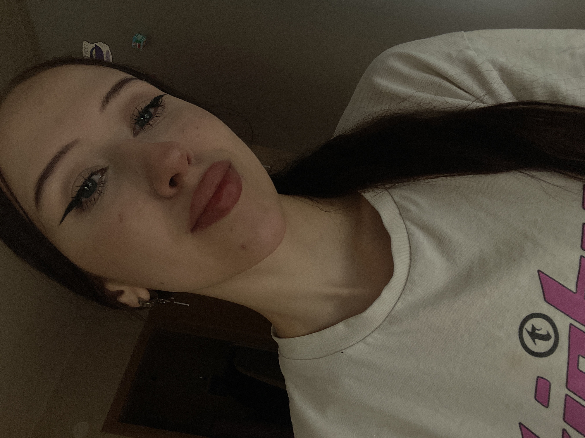 The image is a close-up, dimly lit selfie of a young person in Votkinsk, Russia. The person is wearing a light-colored t-shirt with pink lettering and a circular graphic on the front. They have dark hair pulled back into a ponytail and are wearing eyeliner and lip gloss. A silver earring with a cross charm is visible on their left ear. The background appears to be an indoor setting with a doorframe on the left and a plain wall on the right.