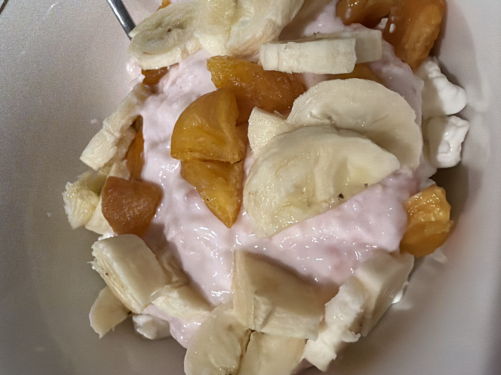 A top-down, close-up shot shows a bowl of food. The bowl appears to be white. Inside, there's pink yogurt with sliced bananas and pieces of dried apricots. The bananas are sliced into rounds, and the apricots are cut into smaller, irregular pieces. Some white cream, possibly cottage cheese or yogurt, can be seen near the top right of the bowl. A silver spoon is also visible. There is no evidence of any location being Chaikovsky, Russia in this image.