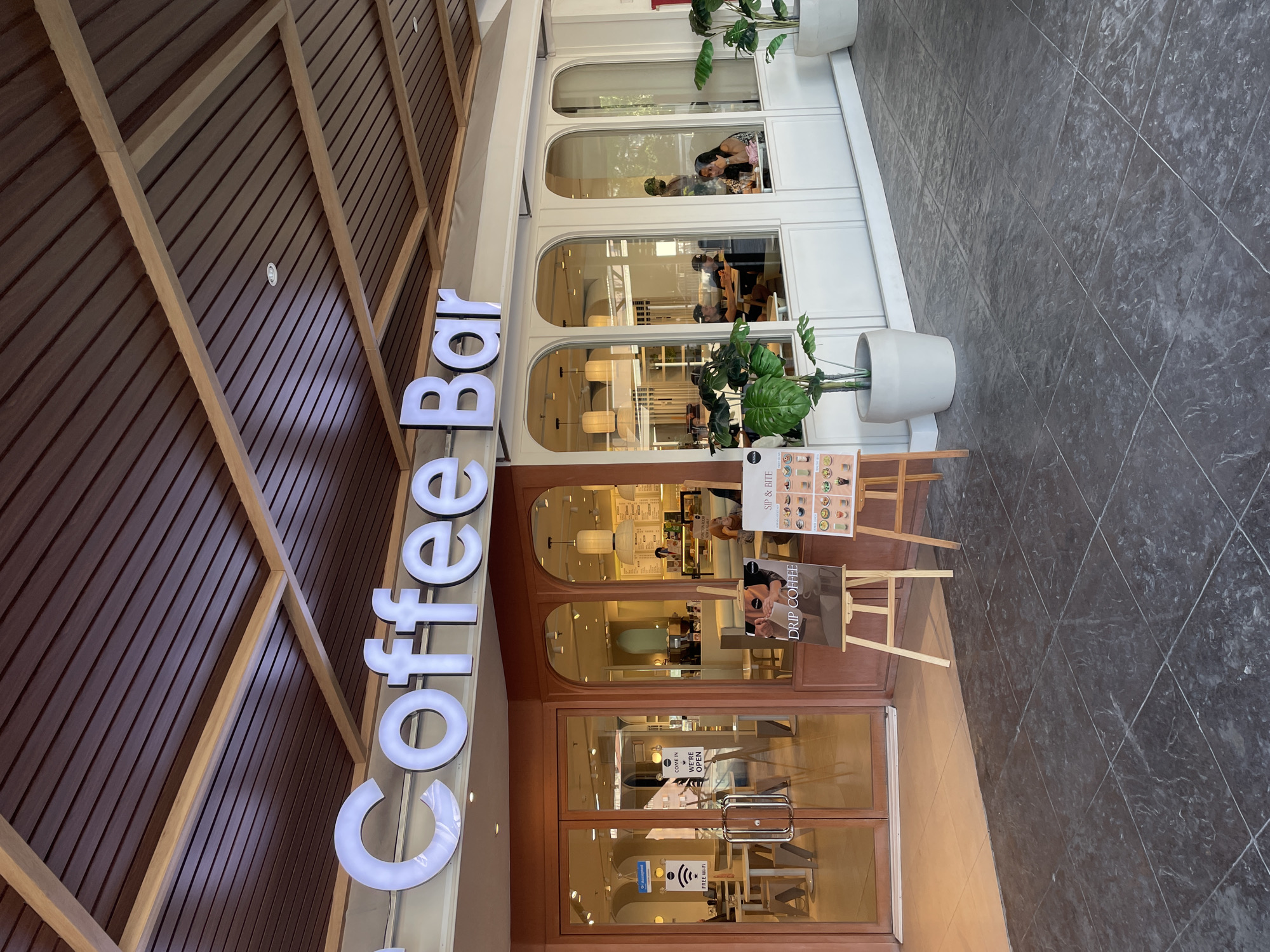 This is a medium shot, eye-level photograph of the exterior of a coffee shop. The coffee shop has a modern, minimalist design with a prominent "Coffee Bar" sign in white neon lettering. The facade is a mix of warm brown and white, with large glass windows revealing the interior. Inside, the seating area is visible, with tables and chairs. Several people are seated inside, enjoying their drinks. A few plants are placed outside the coffee shop, adding a touch of greenery. Two easels with signs advertising "Drip Coffee" and "Sip & Bite" are placed in front of the entrance. The floor in front of the coffee shop is paved with dark grey tiles. The overall atmosphere is relaxed and inviting. The coffee shop is located in Davao City, Philippines.