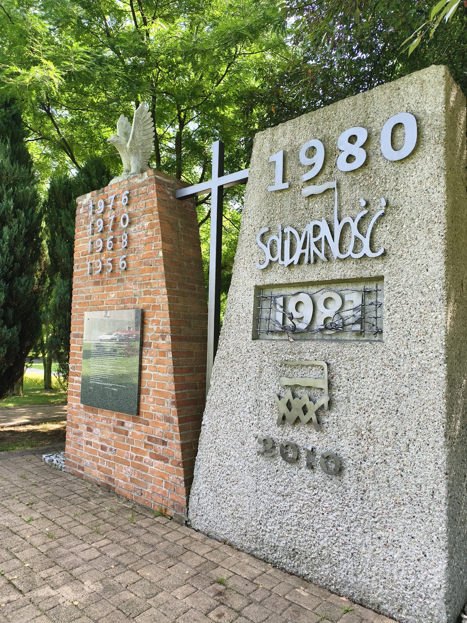 The image shows a monument in Tczew, Poland, dedicated to the Solidarity movement. The monument is composed of two parts: a brick pillar topped with a sculpture of an eagle, and a larger concrete structure. The brick pillar has the years "1976", "1970", "1968", and "1956" inscribed on it, along with a plaque displaying text and an image. The concrete structure features the year "1980" prominently displayed, followed by the word "SOLIDARNOŚĆ" and the year "1981" enclosed within a metal grate. Below this, there is another metal symbol and the year "2010". The monument is situated outdoors, with trees and foliage visible in the background, and a paved walkway in the foreground. The scene is brightly lit, suggesting daytime.