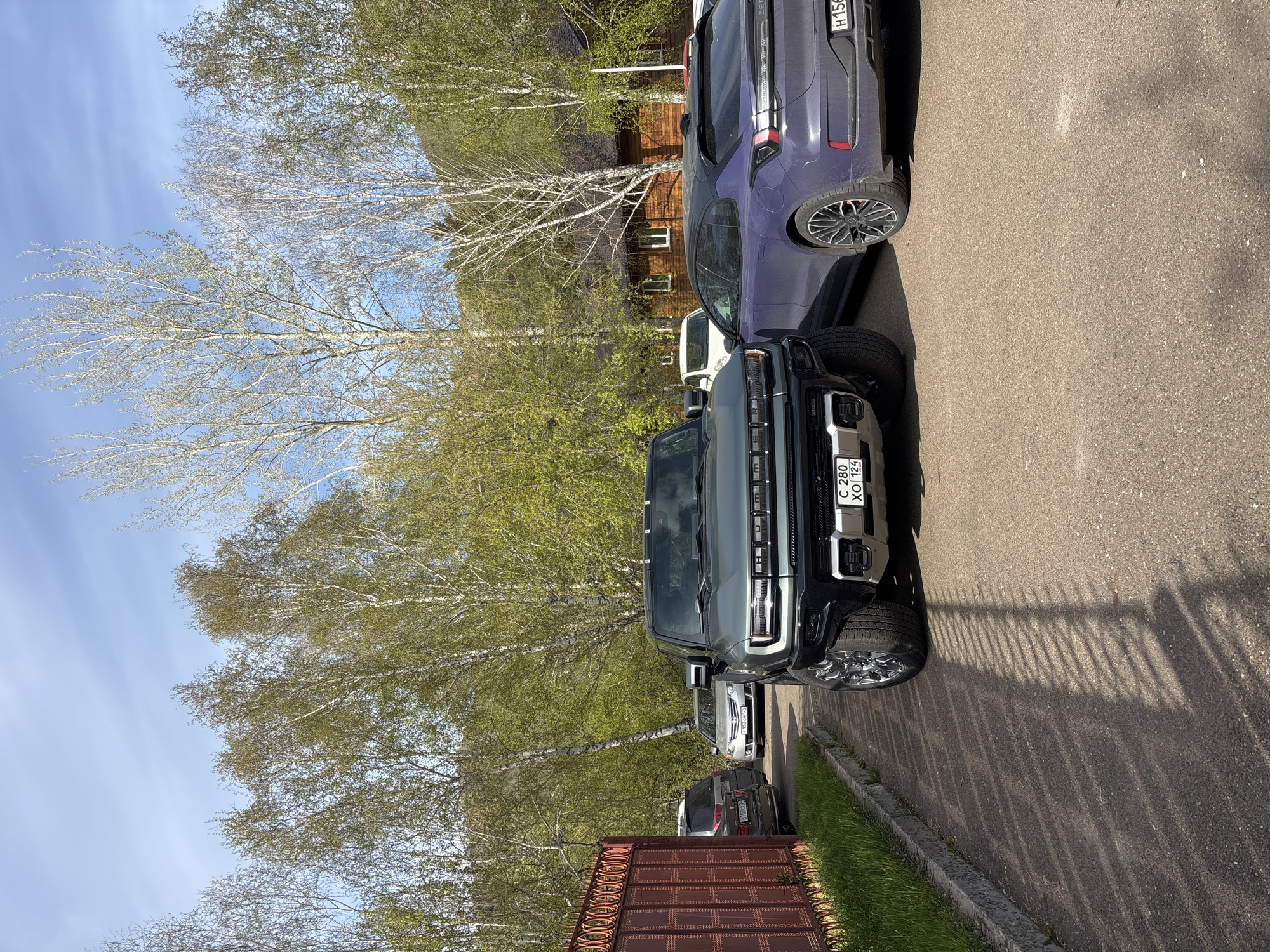 The image shows a street scene in Krasnoyarsk, Russia, with several parked vehicles. A black GMC Hummer EV with a Russian license plate "C 280 XO 124" is prominently featured. Next to it is a dark purple SUV, and further down the street are several other parked cars. The background includes trees with sparse foliage, suggesting early spring or late autumn, and a wooden building. The sky is blue with scattered clouds. A brown metal fence is visible in the lower left corner. The scene is captured in bright daylight, casting long shadows on the asphalt road.