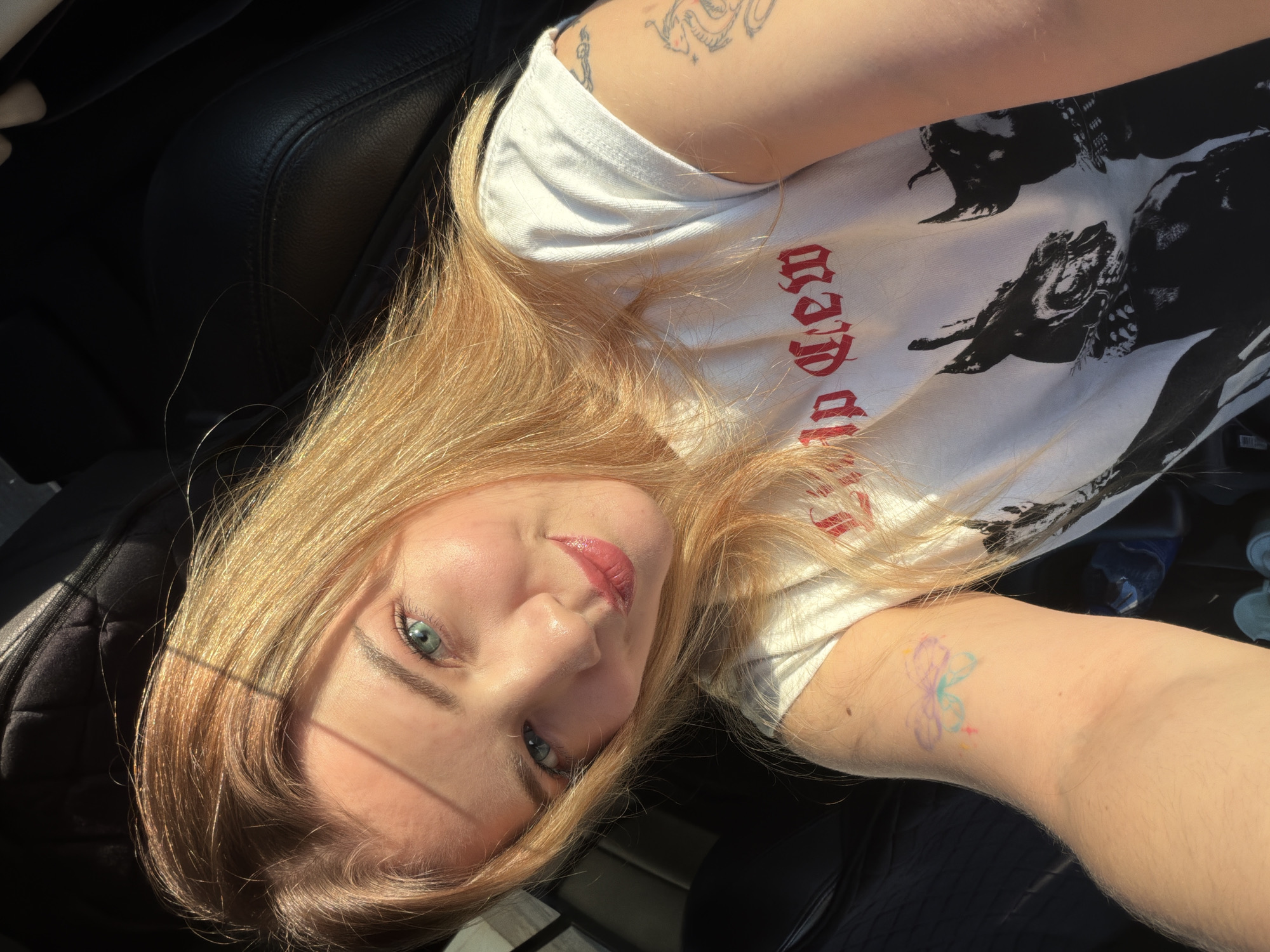 A selfie of a blonde woman with blue eyes, lying in the driver's seat of a car. She is wearing a white t-shirt with a black and white print and red lettering. Tattoos are visible on her arms. The car seat is black leather. The exact location in Naberezhnye Chelny, Russia, is not discernible from the image.