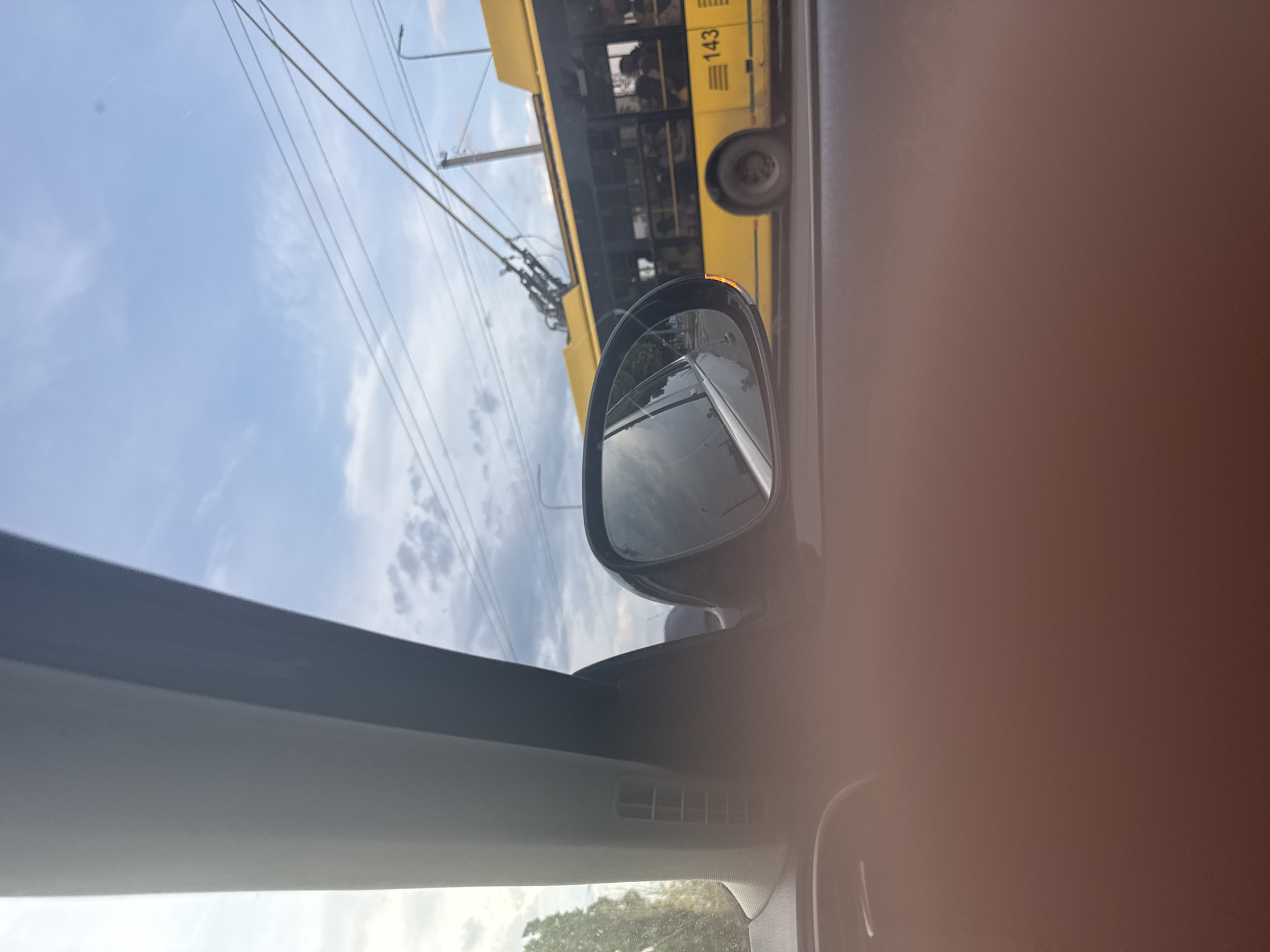 The image captures a street scene from the interior perspective of a vehicle, looking out the passenger-side window. In the immediate foreground, the lower portion of the image is dominated by a blurry, reddish-brown surface, likely a close-up, out-of-focus part of the car's interior or an occupant's limb. Above this, the car's grey A-pillar is visible on the left, and a black side mirror is positioned to its right, reflecting a blurred view of the road and sky.
Outside the vehicle, a bright yellow trolleybus with the route number "143" prominently displayed on its lower right side occupies the midground, moving from left to right. Passengers are visible through its multiple rectangular windows, some seated, others standing. Two trolley poles extend from the bus's roof to connect with overhead electrical lines (catenary wires) that crisscross the sky. A utility pole stands behind the trolleybus, also supporting these lines. To the far left, beyond the A-pillar, blurred green foliage from trees is visible against the bright sky. The background consists of a partly cloudy blue sky with scattered white and grey clouds. The scene depicts an urban or suburban street in the city of Unknown, Unknown.