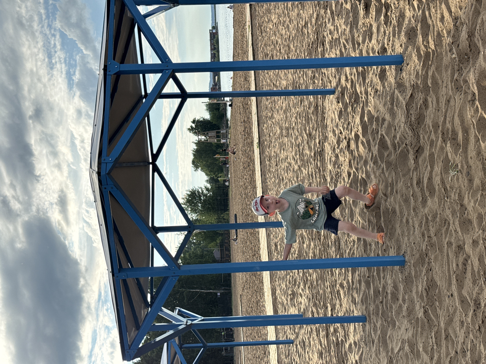 A young boy with fair skin, wearing a green t-shirt with a "CAMPING" graphic, dark shorts, a red and white baseball cap, and orange sandals, stands inside a blue, octagonal beach shelter on a sandy beach. His mouth is open and his eyes are wide as he looks towards the left, with his left arm extended. The beach shelter has a metallic frame with a grey fabric roof. In the background, there is a calm body of water, trees, and buildings across the water. The sky is cloudy. The scene appears to be a public beach area, possibly within the city of Chaikovsky, Russia, given the context.