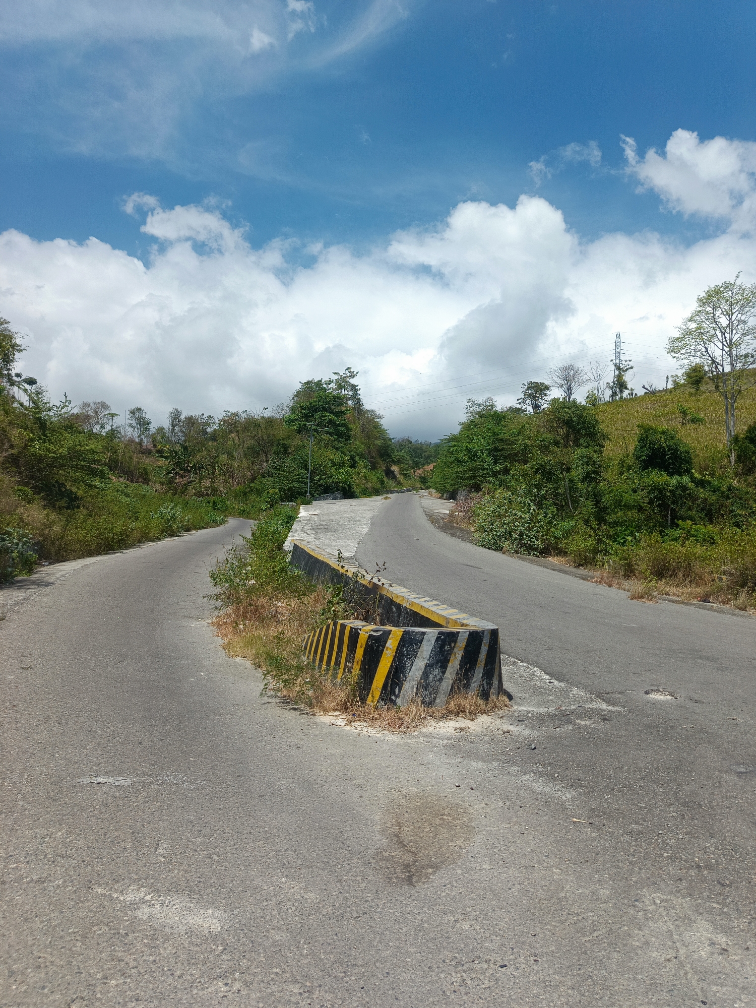 The image depicts a rural road in the city of Ilotidea, Indonesia, under a bright, partly cloudy sky. The road forks, with one path curving to the left and the other to the right, both leading uphill into a vegetated landscape. A concrete barrier with black and yellow diagonal stripes is positioned at the fork, marking a separation between the two road sections. The surrounding environment is characterized by lush green trees and vegetation, with some dry grass and sparse foliage visible along the roadside. In the distance, a transmission tower and power lines are discernible against the hilly terrain. The road surface appears to be asphalt, showing signs of wear and tear with some patches and cracks. No people or animals are present in the scene. The overall impression is of a remote, undeveloped area.