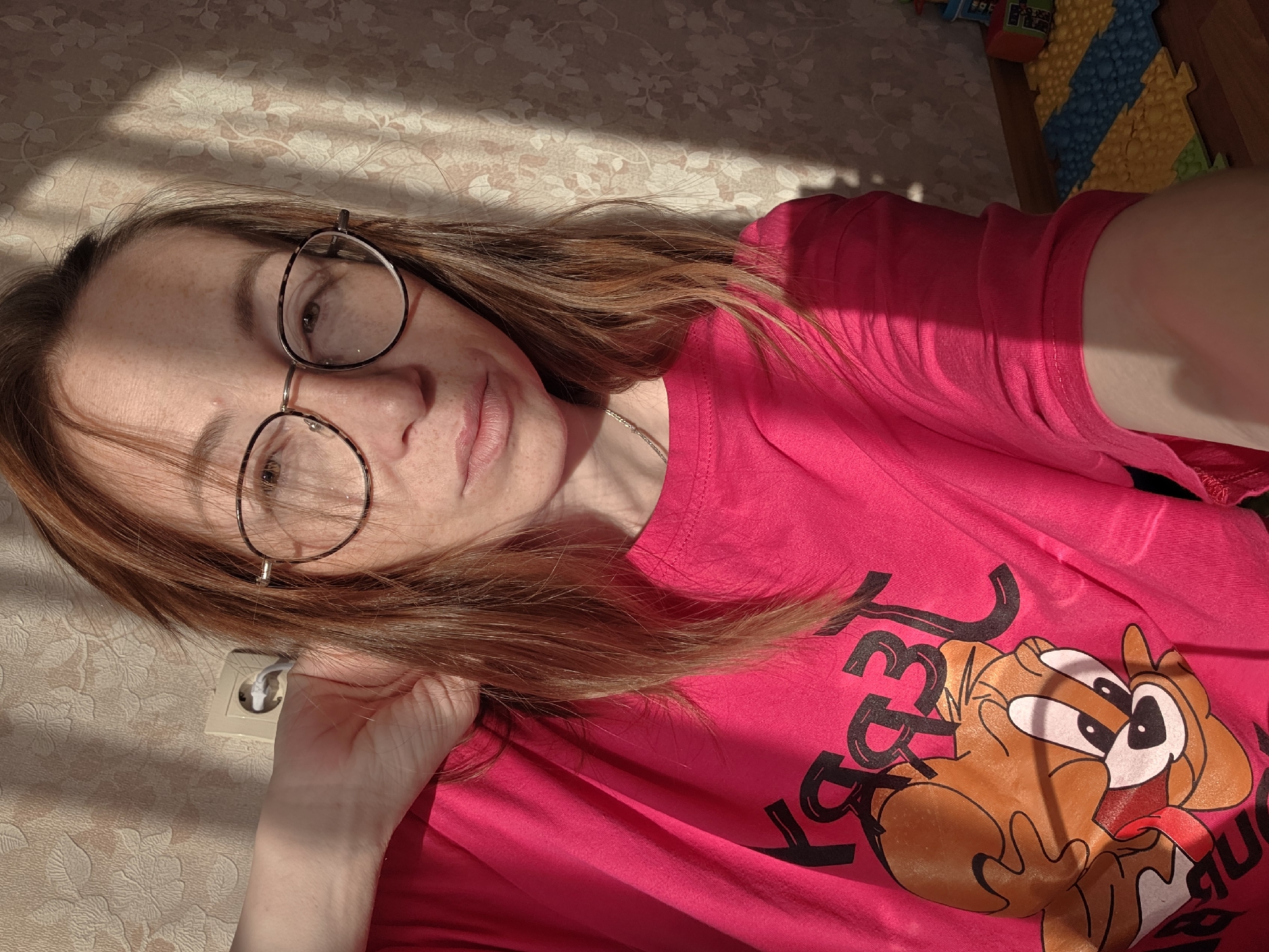 A person is lying down in a brightly lit room, possibly taking a selfie. The person has fair skin, light brown hair, and is wearing round glasses. They are wearing a pink t-shirt with a cartoon character print. The background includes a textured wall with sunlight casting patterned shadows, and a glimpse of a colorful children's play mat. An electrical outlet is visible on the wall near the person's hand.