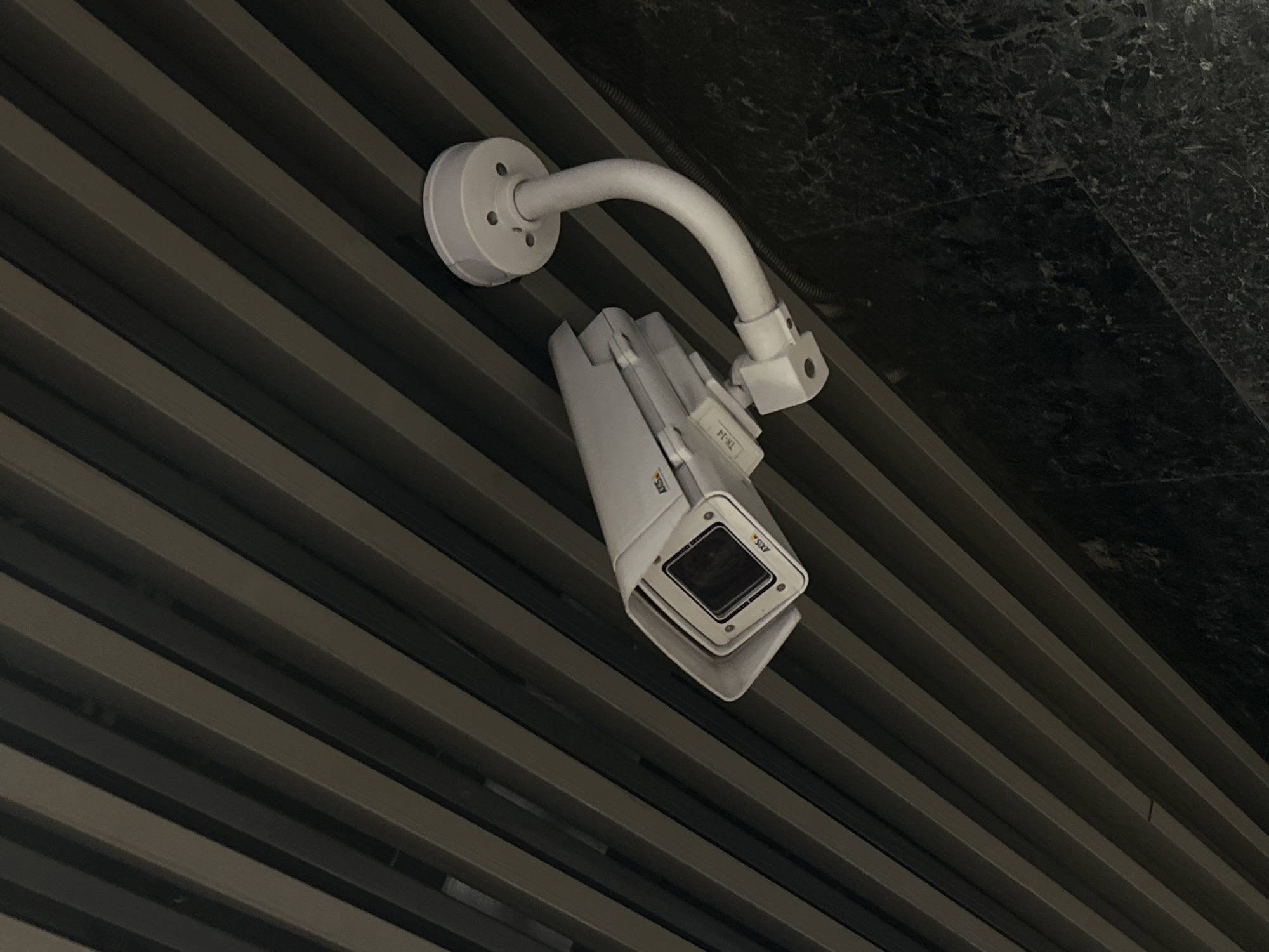 A white security camera is mounted on a white curved arm, affixed to a ceiling consisting of parallel, gray-toned linear panels. A section of the ceiling transitions to a dark, textured surface, possibly stone or heavily patterned wallpaper. The camera is labeled "AXIS." The location is not identifiable from the image.