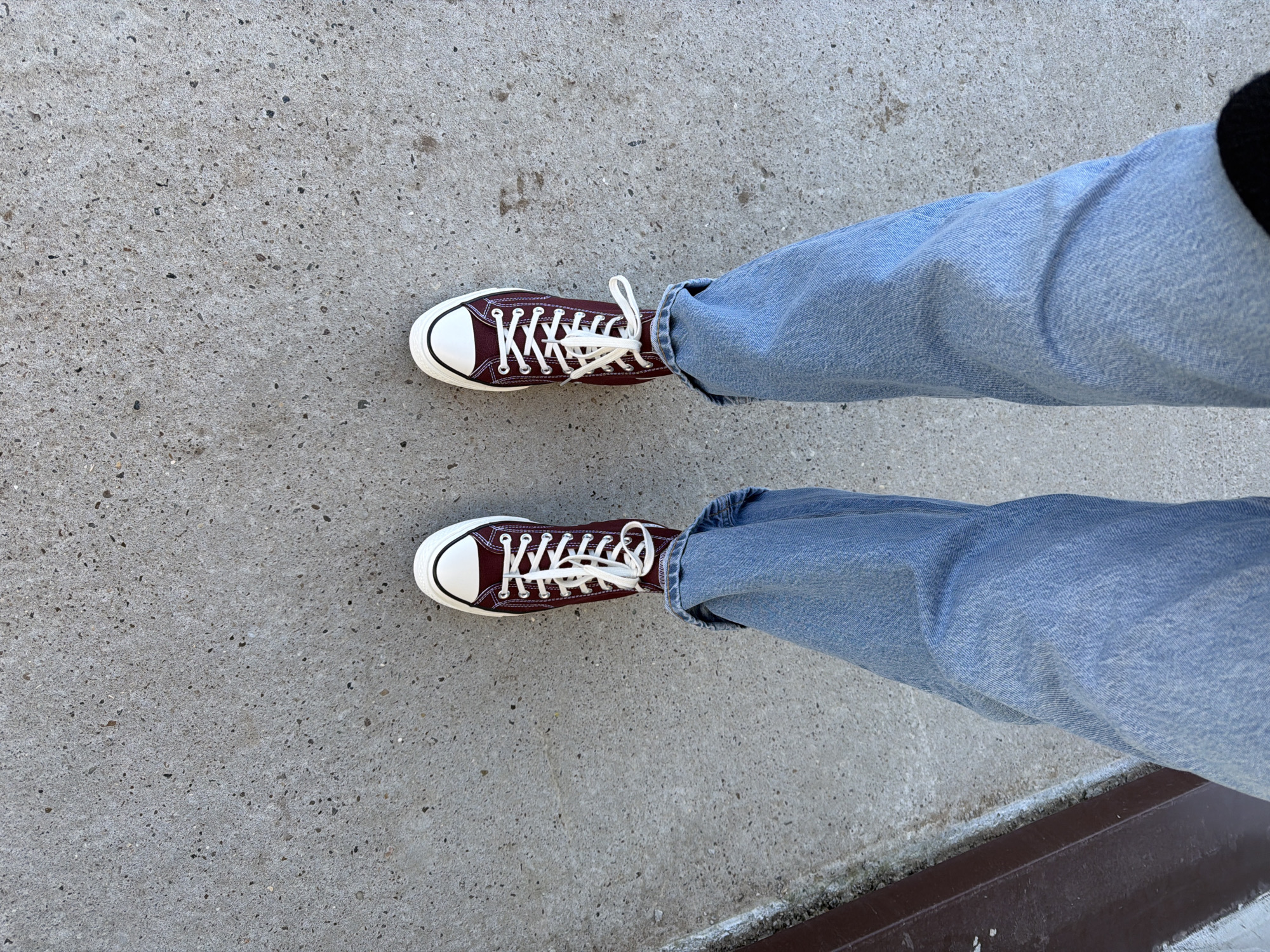 The image presents an overhead view of a person's legs and feet, likely standing on an outdoor surface. The person is wearing light blue denim jeans that are casually cuffed or pooled around the ankles. On their feet are a pair of high-top sneakers, distinctive with their dark maroon or burgundy canvas uppers, white rubber toe caps, and bright white laces tied in bows. The soles of the sneakers are also white, contrasting with the dark body.
The setting is a textured grey concrete surface, appearing to be pavement or a walkway, speckled with small dark and light aggregate. Along the very bottom edge of the frame, a dark, linear object, possibly a curb or metal beam, is visible, with some lighter residue or dirt beside it. The lighting suggests it is daytime, with clear, even illumination. There is no visible text in the image. According to the provided context, the location is Chaikovsky, Russia.