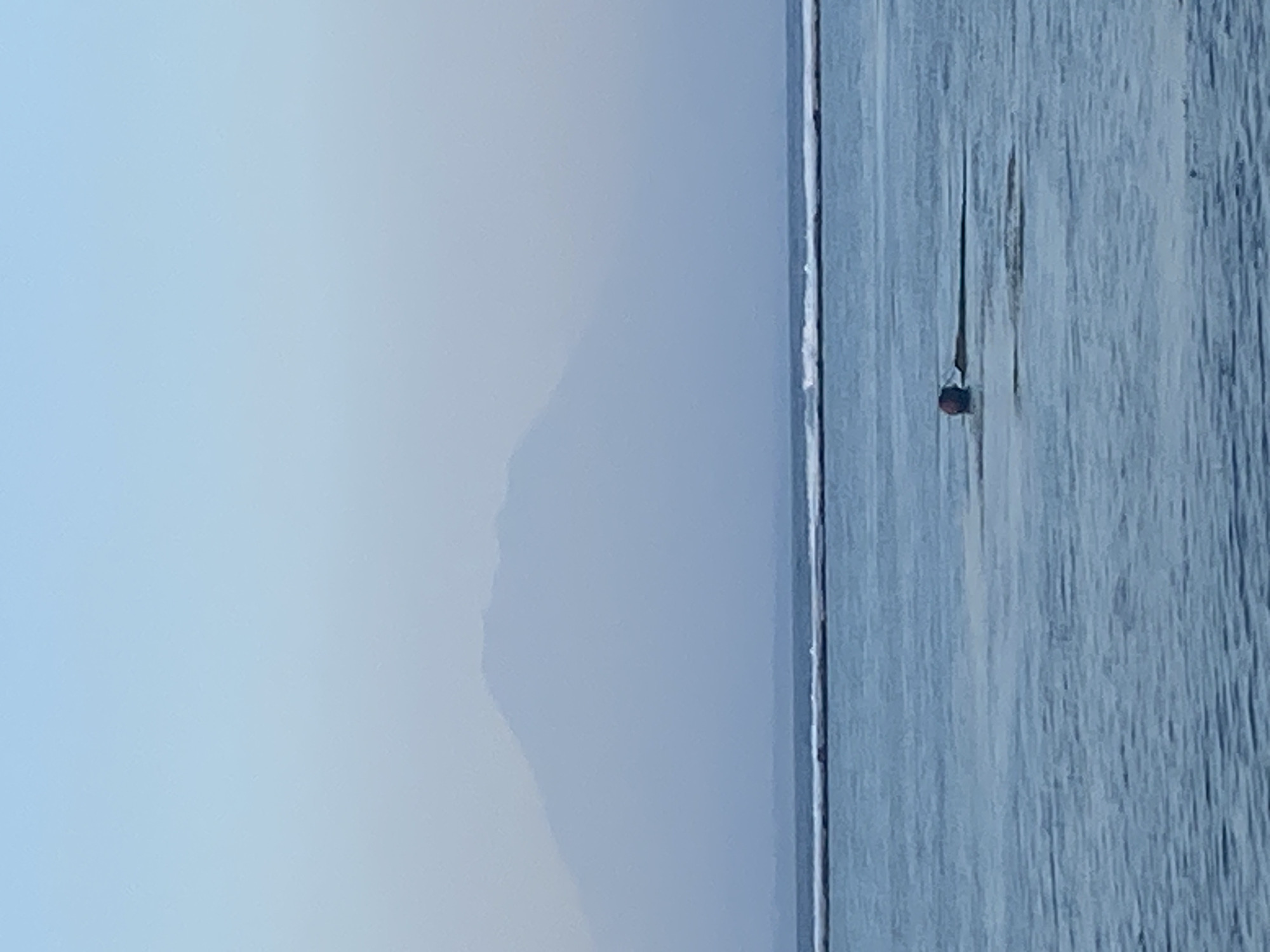The image depicts a serene seascape with a distant, hazy mountain range under a pale blue sky. In the foreground, calm, blue water is visible with subtle ripples. A thin line of white surf marks the breaking waves in the mid-ground. Near the center of the frame, a red buoy is partially submerged in the water, attached to a dark, elongated object that appears to be floating. The overall atmosphere is tranquil and expansive. This scene could be observed from a coastal area in Denpasar, Indonesia, such as the beaches along the southern coast, where Mount Agung or other inland mountains are often visible in the distance.