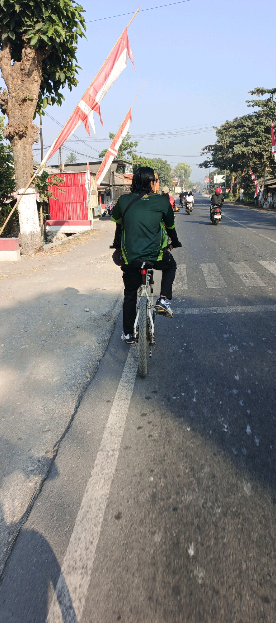 A person rides a bicycle on a paved road in Mlokorejo, Indonesia. The cyclist, viewed from behind, wears a green shirt with yellow accents and black pants. They are on a white bicycle with black tires and silver spokes. The road is marked with a white dashed line on the left side, separating it from a dirt shoulder. In the background, several motorcycles are also present on the road. Buildings with red corrugated metal siding are visible on the left side of the road, adjacent to the dirt shoulder. Red and white flags, possibly national flags, are displayed on poles along the roadside. The sky is clear and blue, indicating a daytime scene. The overall environment suggests a moderately trafficked street in a town or village setting.