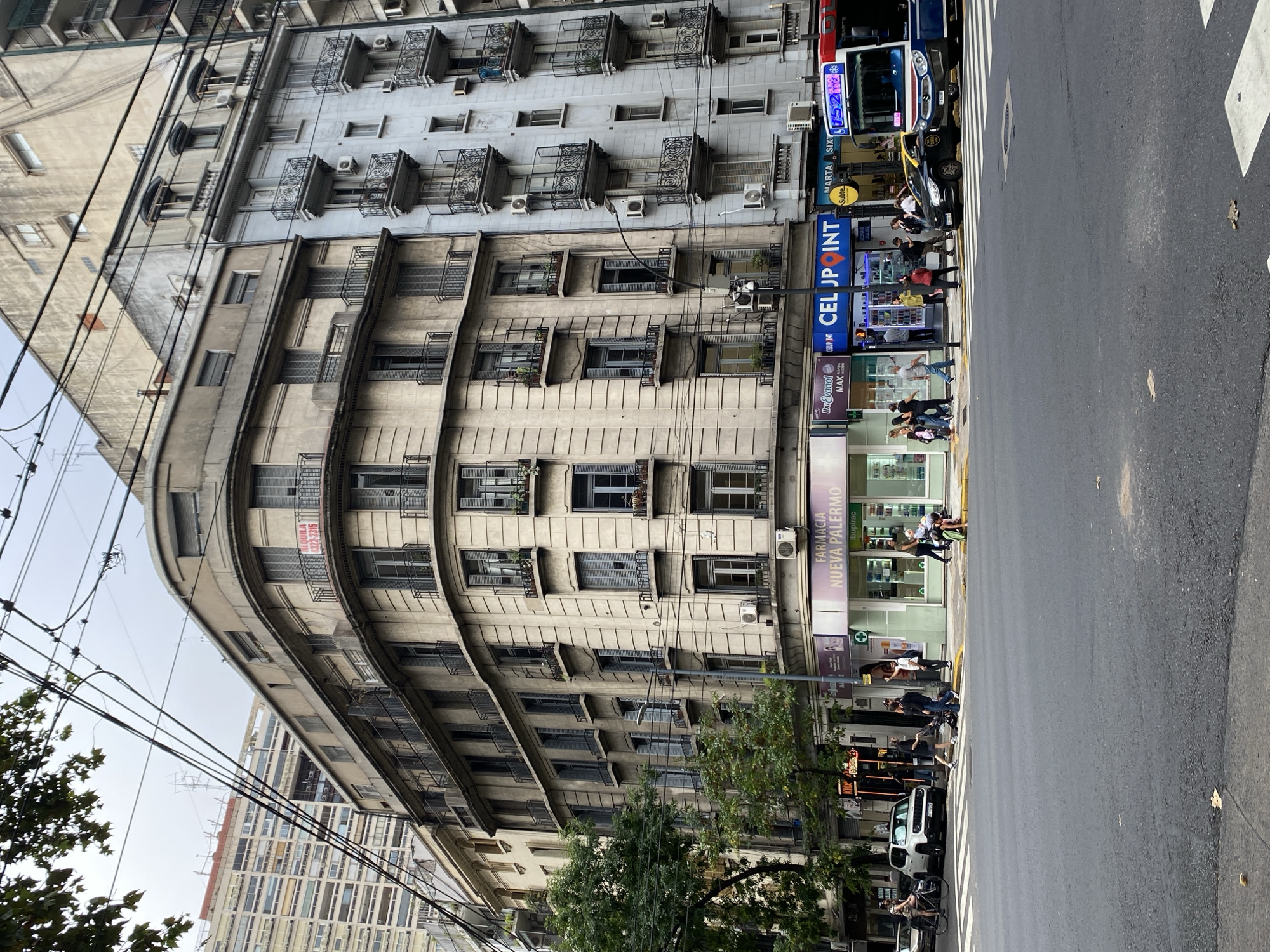 The image depicts a daytime street scene at an intersection in the Palermo neighborhood of Buenos Aires, Argentina, under an overcast sky. The foreground features an asphalt road with white crosswalk lines and scattered dry leaves.
The dominant structure is a multi-story, beige-toned corner building with a rounded facade, featuring numerous windows with light blue shutters and balconies with dark, ornate railings. A red "ALQUILA" (For Rent) sign with a phone number (4322-2315) is visible on a third-floor balcony. The ground floor houses "FARMACIA NUEVA PALERMO" with a green cross logo and "CELU POINT" (a mobile phone shop) with bright blue signage. Adjacent to the corner building on the right is a taller, light blue/grey building with similar balconies and multiple external air conditioning units. Further left, behind trees and utility wires, a modern, light-colored building with many windows is partially visible. Numerous black utility wires crisscross the upper portion of the frame. A black street lamp post and a yellow "Subte" (subway) sign are present on the sidewalk.
Approximately 20-25 individuals are observed. On the left side of the street, a person on a bicycle is near a white Jeep Renegade. Several pedestrians are walking on the sidewalks and crossing the street. One individual in a black shirt is bent over near the crosswalk, seemingly interacting with an object on the ground, while another person stands nearby. On the right sidewalk, a woman in a red top is crossing the street, and other pedestrians are walking.
Vehicles include a red and blue city bus (Line 152) stopped on the right side of the road, a black taxi with a yellow roof positioned behind it, and the white Jeep Renegade on the left.
