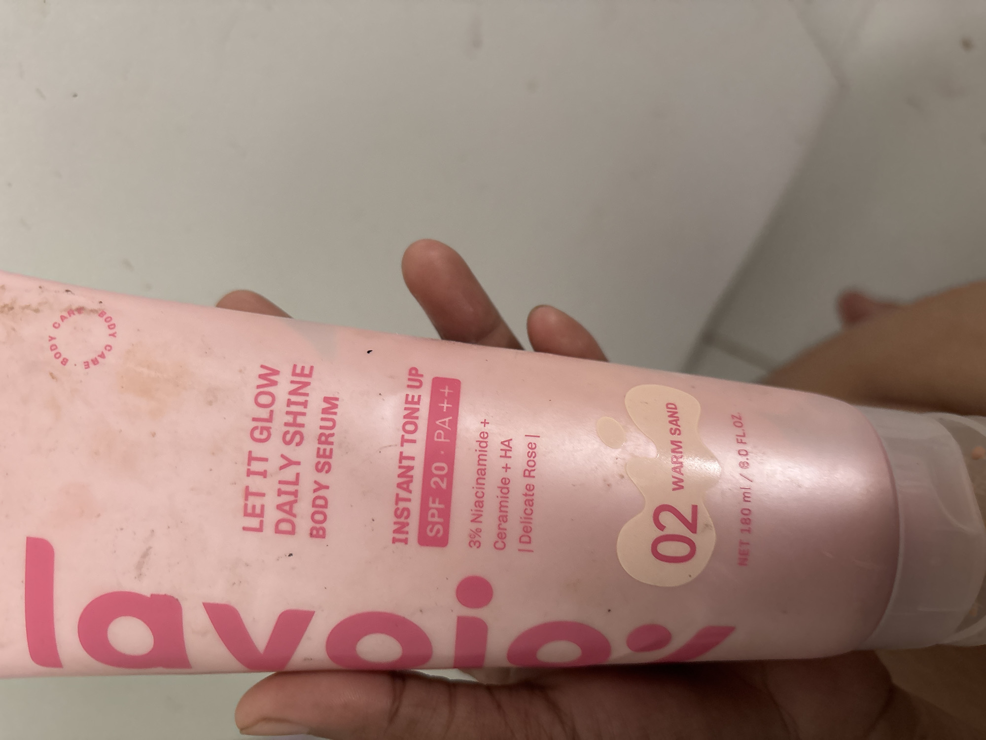 A close-up image shows a pink tube of "lavojoy" brand "Let It Glow Daily Shine Body Serum," specifically the "Instant Tone Up" variant. The product has SPF 20 PA++ and contains ingredients such as 3% Niacinamide, Ceramide, and HA, with a delicate rose scent. The tube indicates the shade as "02 Warm Sand" and contains a net volume of 180 ml / 8.0 FL.OZ. A person's fingers are partially visible, holding the tube. The background is a light-colored surface. The image was likely taken in North Jakarta, Indonesia (based on the brand's origin).