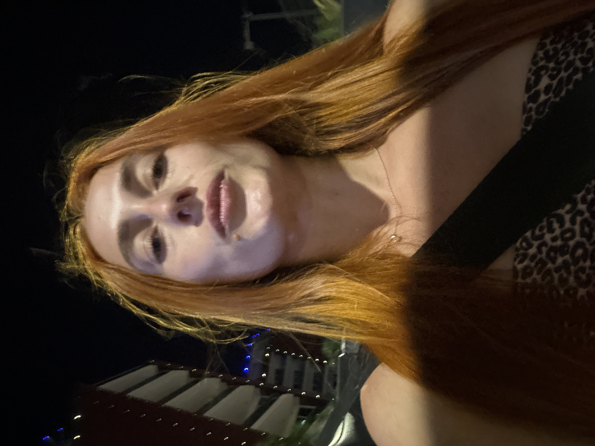 A young woman with long, auburn hair is captured in a selfie at night in Budva, Montenegro. The woman is looking directly into the camera, with her mouth slightly parted. She is wearing a strapless top with a leopard print pattern. In the background, a modern building with balconies and lights is visible, along with palm trees and a dark night sky. The overall setting suggests an urban environment at night, possibly near a resort or entertainment area.