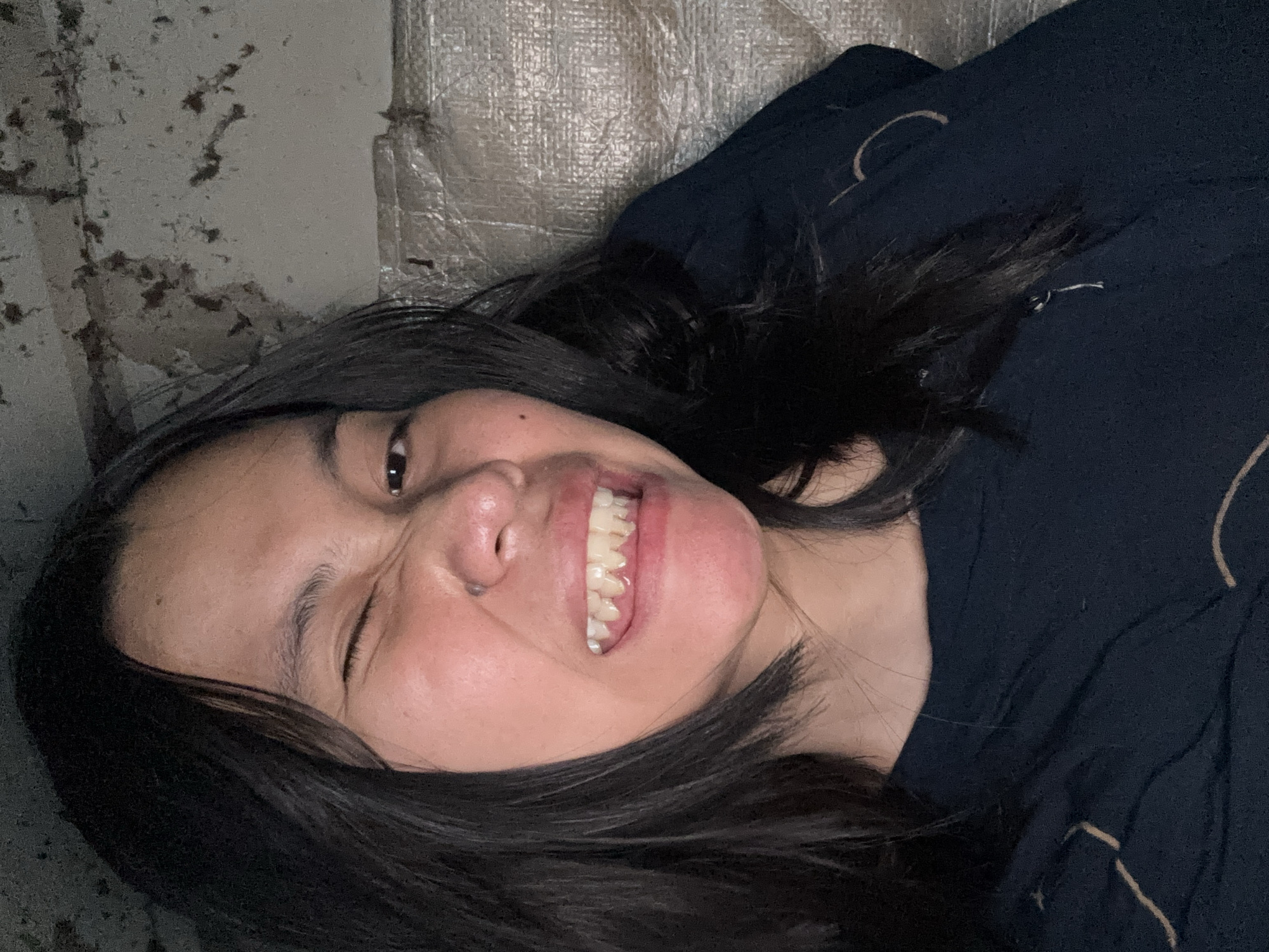 In East Jakarta, Indonesia, a close-up, waist-up, eye-level, indoor shot features a young person with dark, shoulder-length hair. They are wearing a dark blue t-shirt with a faint outline design. The person is winking with their right eye, while their mouth is open, revealing their teeth in a grimace. Their left eye is partially visible, and their skin appears light-toned. The background is dimly lit and textured, with a rough, mottled wall on the left and a silvery, reflective material on the right.