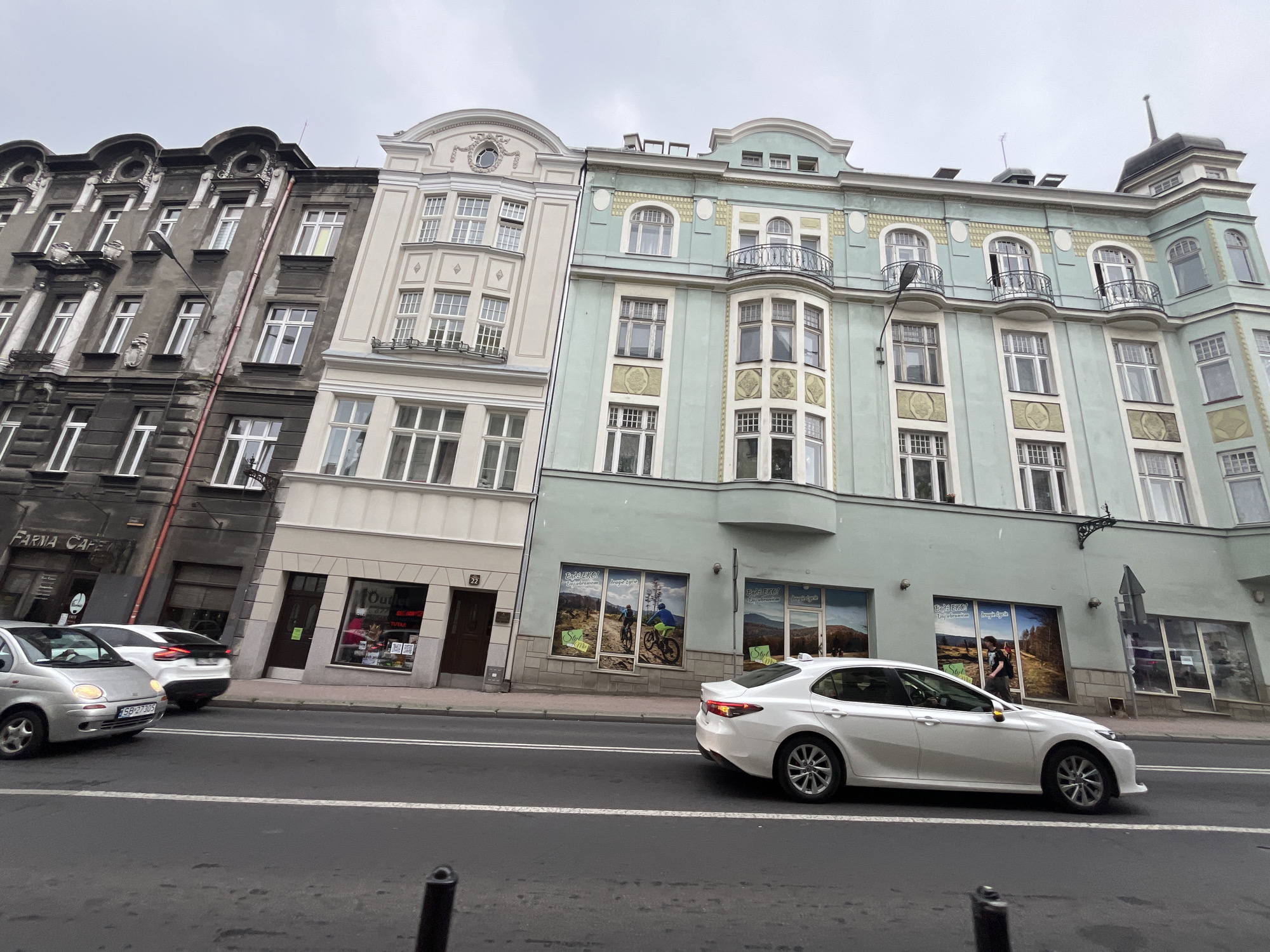 The image shows a street scene in a city with several buildings and cars. The buildings are multi-story, with varying architectural styles. One building is a dark, older structure with ornate detailing. Next to it is a lighter-colored building with a more classical facade. Further to the right is a pale green building with balconies and decorative elements. The ground floor of the green building appears to house businesses, with large windows displaying graphics.
In the foreground, a white sedan is driving on a paved road. A silver hatchback is also visible on the road. The sky is overcast. Streetlights and a traffic signal are present. The environment appears to be a commercial or mixed-use area.