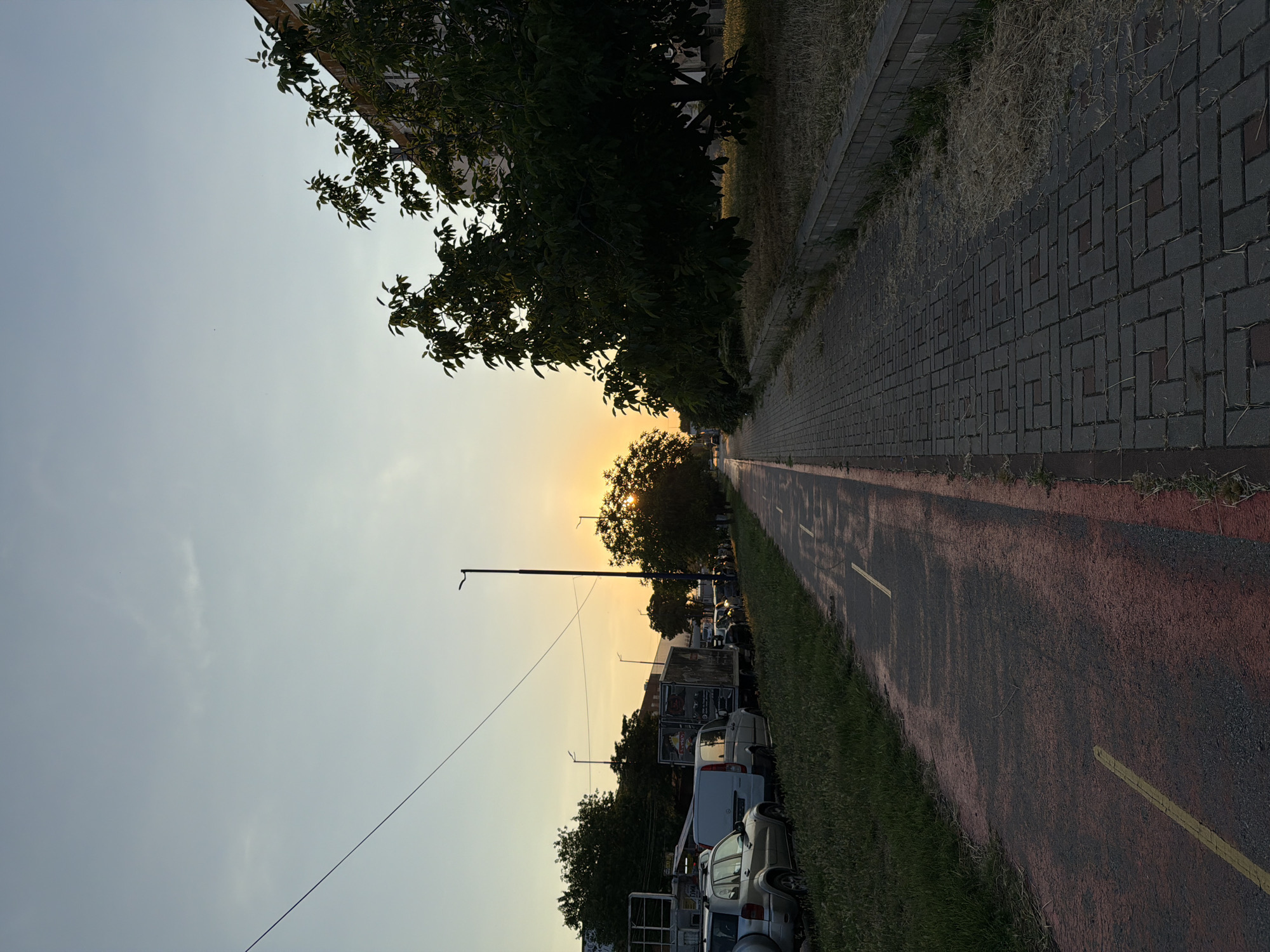 This is a photograph taken at sunset in Pazardzhik, Bulgaria. The image shows a paved pedestrian walkway alongside a bike lane on the right and a grassy area with parked cars and buildings on the left. The sun is low in the sky, casting a warm glow and creating long shadows. The scene appears to be a typical urban environment, possibly a residential area or a commercial district. There are no people visible in the image, and the focus is on the landscape and architecture. The overall atmosphere is peaceful and serene, with the natural beauty of the sunset contrasting with the man-made structures.