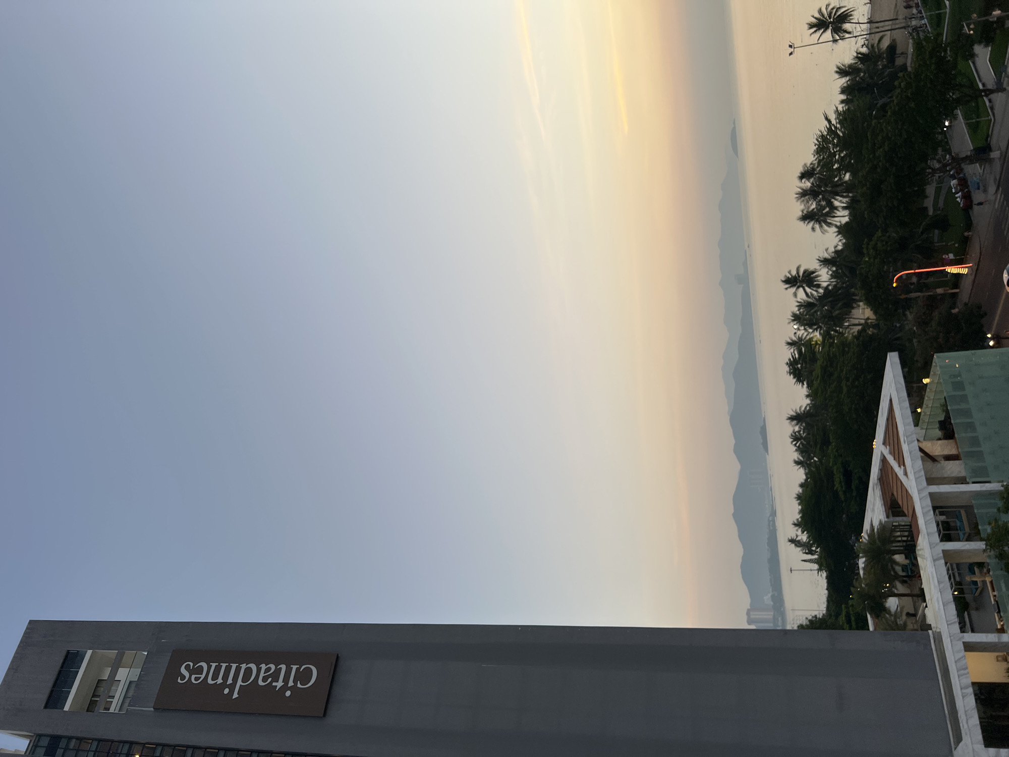The image shows a view from a building in Nha Trang, Vietnam, at dusk. On the left is a tall, gray building with a sign that reads "citadines". In the distance, the sea is visible with a hazy coastline and mountains. Palm trees and lush green foliage line the beachfront. The sky is a soft gradient of blue and pale orange, indicating the setting sun. The scene appears peaceful, with a few streetlights illuminating the area near the beach.