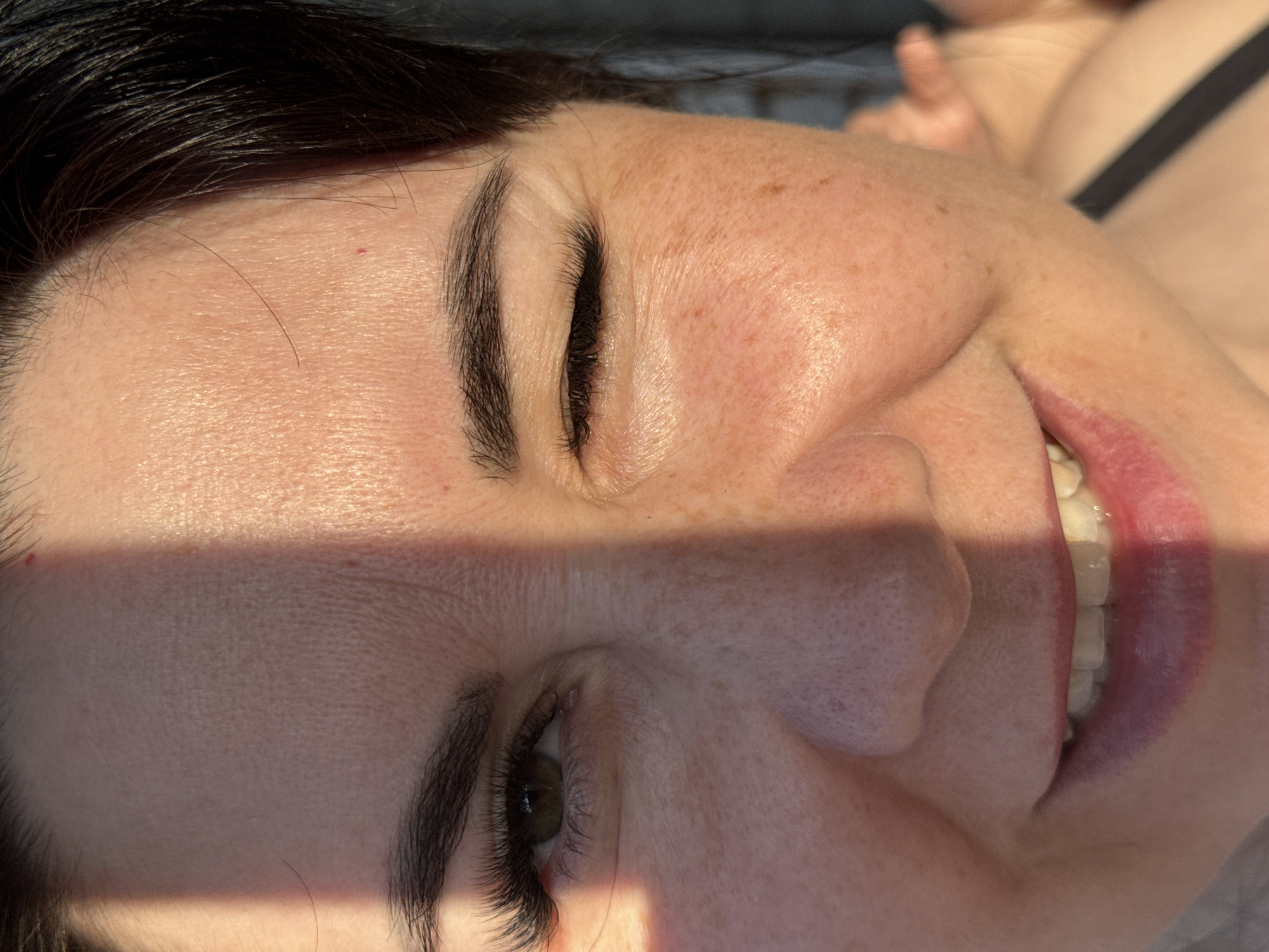 A close-up, partially shadowed view of a woman's face. She has dark hair, fair skin with freckles, and dark eyelashes. Her lips are visible, showing white teeth. Part of her shoulder and the strap of her clothing are visible in the background. The image does not contain information to determine a specific location.