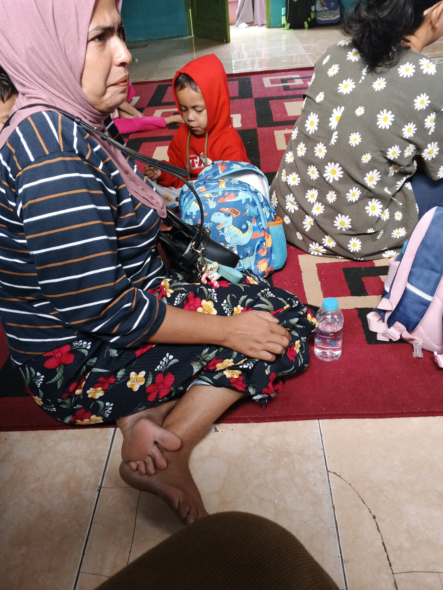 In Warungkiara, Indonesia, several people are seated on a red carpet on the floor. In the foreground, a woman wearing a pink hijab and a striped long-sleeved shirt is visible from the chest down. She has on a floral skirt with red and yellow flowers. Her bare feet are crossed in front of her. To her right, a young child in a red hooded sweatshirt is looking down. A blue backpack with a dinosaur print is placed next to the child. Behind the child, another person is seated, wearing a dark green shirt with a pattern of white daisies. To the right, a pink and blue backpack is partially visible. A clear plastic bottle of water with a blue cap sits on the carpet near the woman's skirt. The environment appears to be indoors, with tiled flooring visible to the left of the carpeted area and what looks like a doorway or wall in the background.