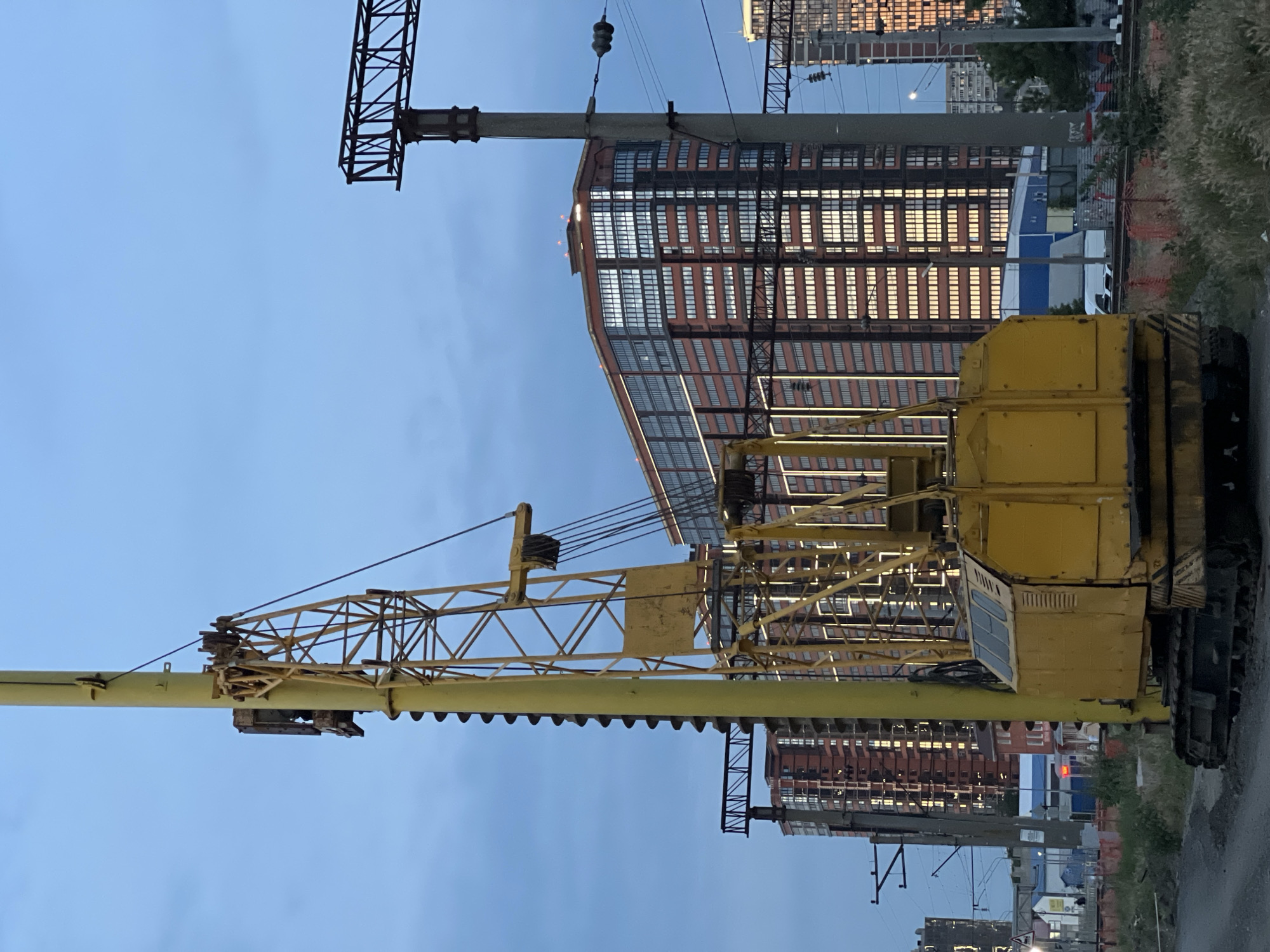 A yellow crane with a tall mast and a spiral drill bit is positioned in the foreground of a city scene at dusk. Behind the crane, a modern residential building with a facade of red brick and numerous reflective windows stands prominently. To the right of the building, a concrete utility pole with electrical wires and insulators is visible. The sky is a dusky blue, with some light clouds. The ground in the foreground is rough, with gravel and sparse vegetation. In the background, other buildings and structures can be seen, along with what appears to be railway tracks and overhead power lines, suggesting an urban or industrial setting near transportation infrastructure.