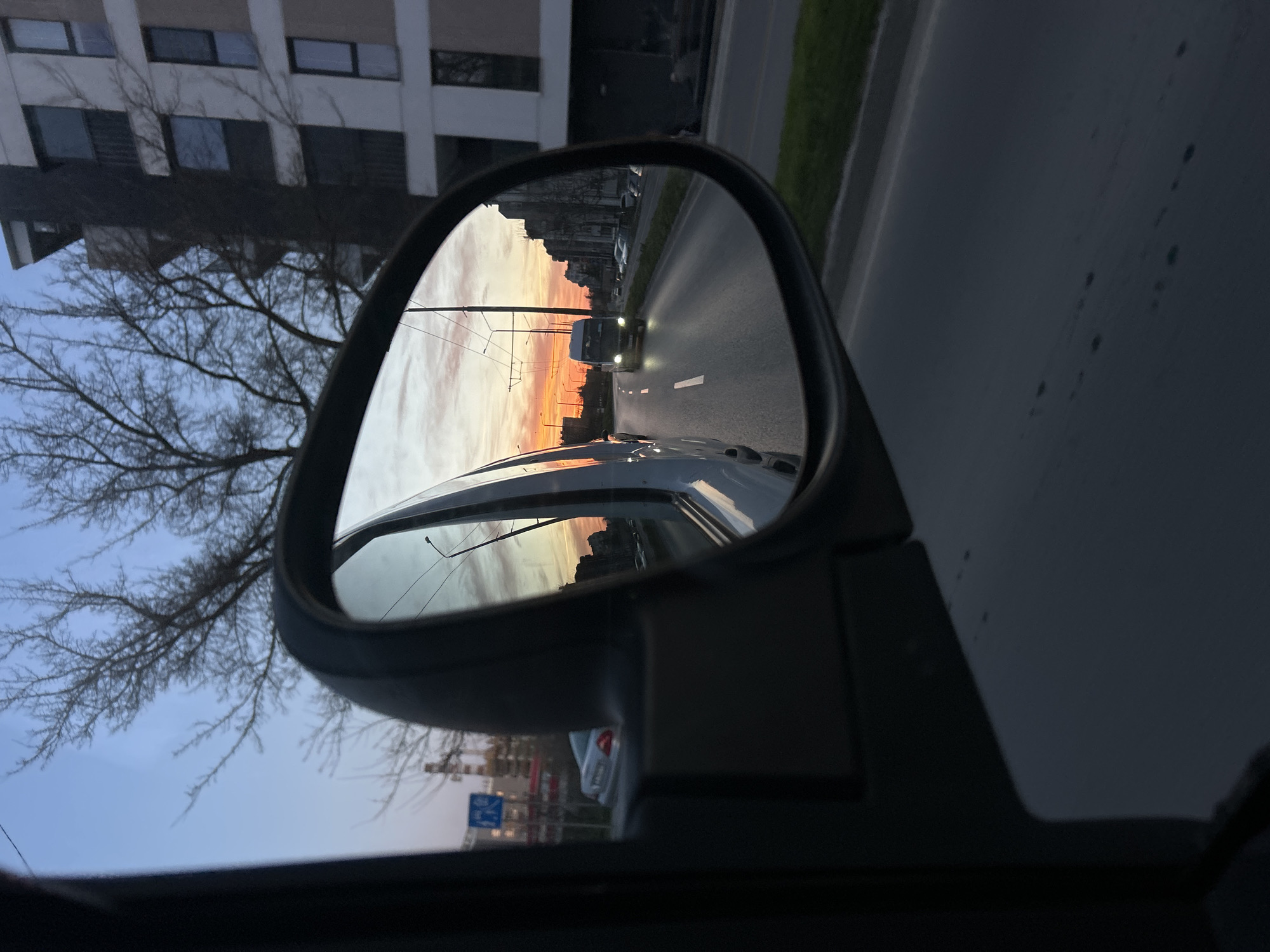 The image depicts a view from a car's side mirror, reflecting a sunset over a road in Varna, Bulgaria. A large truck with illuminated headlights is visible in the mirror, traveling on the road. The sunset colors are reflected in the sky and on the buildings lining the road. Outside the mirror, there are buildings with windows and a leafless tree against a blue sky. Part of the car's interior and side are visible. A road sign is partially visible in the lower left.