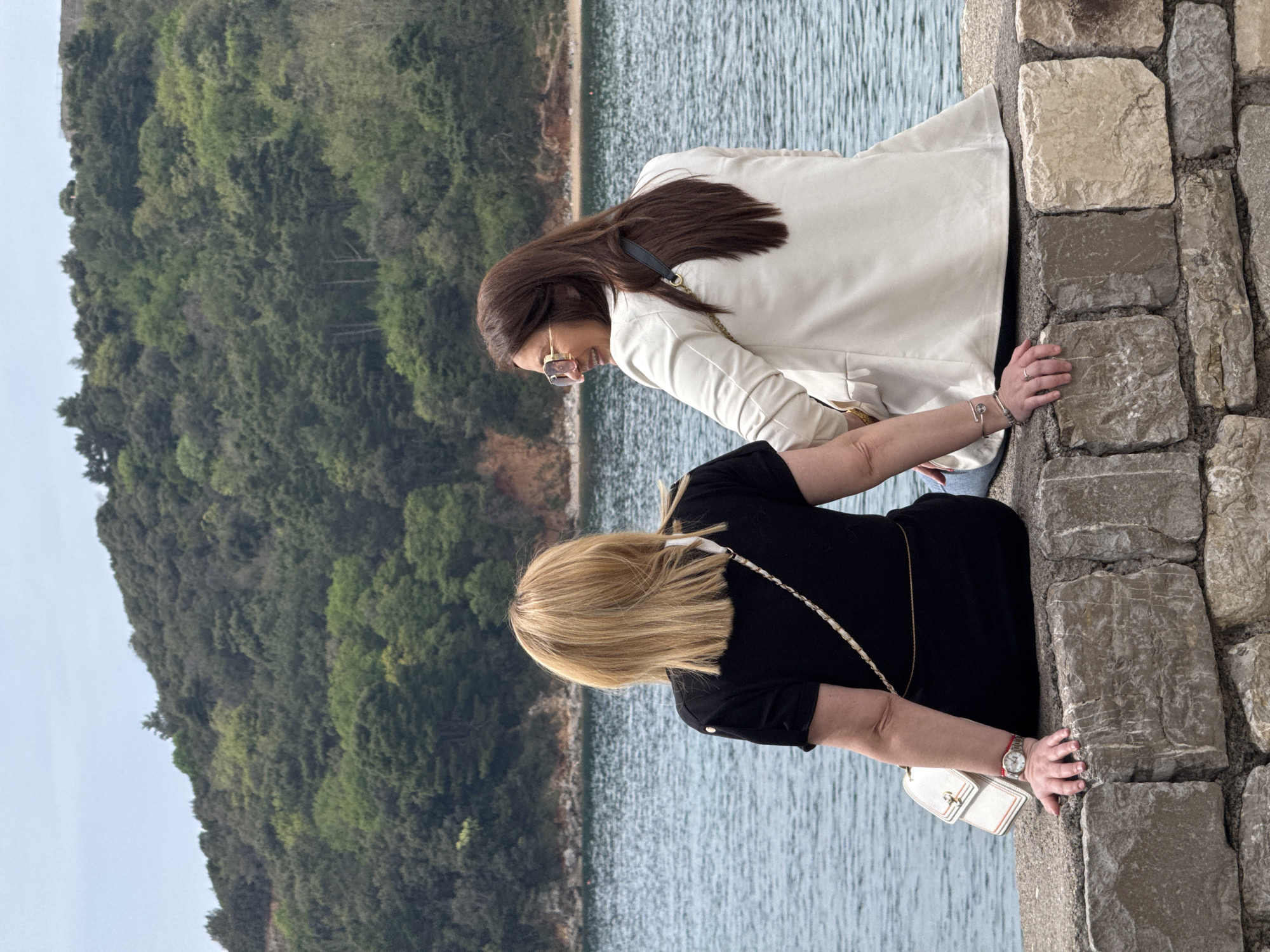 Two women stand side-by-side on a stone wall, overlooking the Adriatic Sea and a tree-covered coastline in Budva, Montenegro. The woman closest to the viewer has blonde hair and wears a black dress with a white purse hanging from her shoulder. She rests her hands on the stone wall. The other woman has long brown hair and wears a white blazer and sunglasses. She is also resting her hands on the wall. The stone wall is made of large, unevenly shaped stones. The sea is in the background.