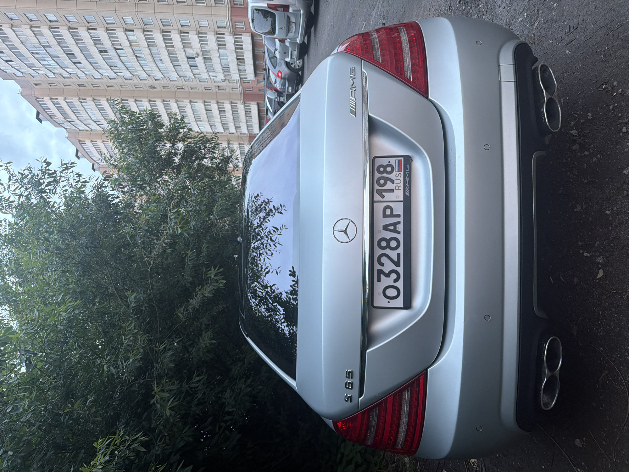 A silver Mercedes-Benz S65 AMG sedan is parked on asphalt in front of a large, modern apartment building. The car's rear is facing the camera, showcasing its taillights, chrome trim, and dual exhaust pipes. A Russian license plate reading "O328AP 198 RUS" is visible. The background includes lush green trees to the left and multiple parked cars, including a white minivan, to the right. The sky is overcast.