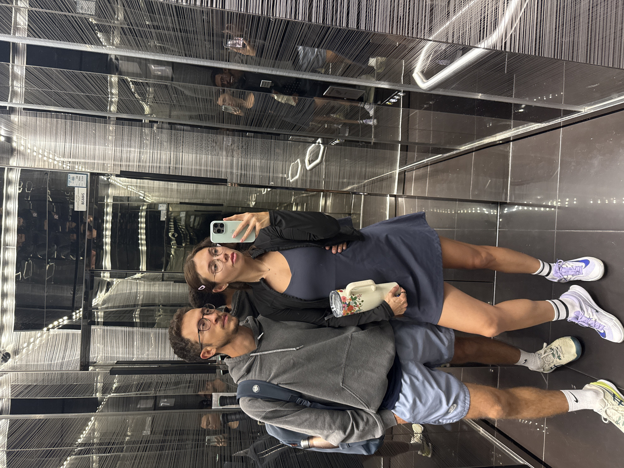 A young adult female and a young adult male are standing in an elevator, taking a mirror selfie. The female is on the right, wearing glasses, a dark navy blue dress, a black jacket, and white and purple running shoes with striped socks. She's holding a floral-designed insulated cup in her right hand and a cellphone in her left, taking the picture. The male is on the left, wearing glasses, a grey hoodie, light blue shorts, and white running shoes with white socks. He has a blue backpack on. The elevator's interior is decorated with vertical lines that could be threads or a design, and it has mirrored surfaces, creating multiple reflections of the couple. The lighting appears to be bright and artificial. Given the artistic design and modern aesthetic of the elevator, it is likely located in a modern hotel or office building in Tel-Aviv.