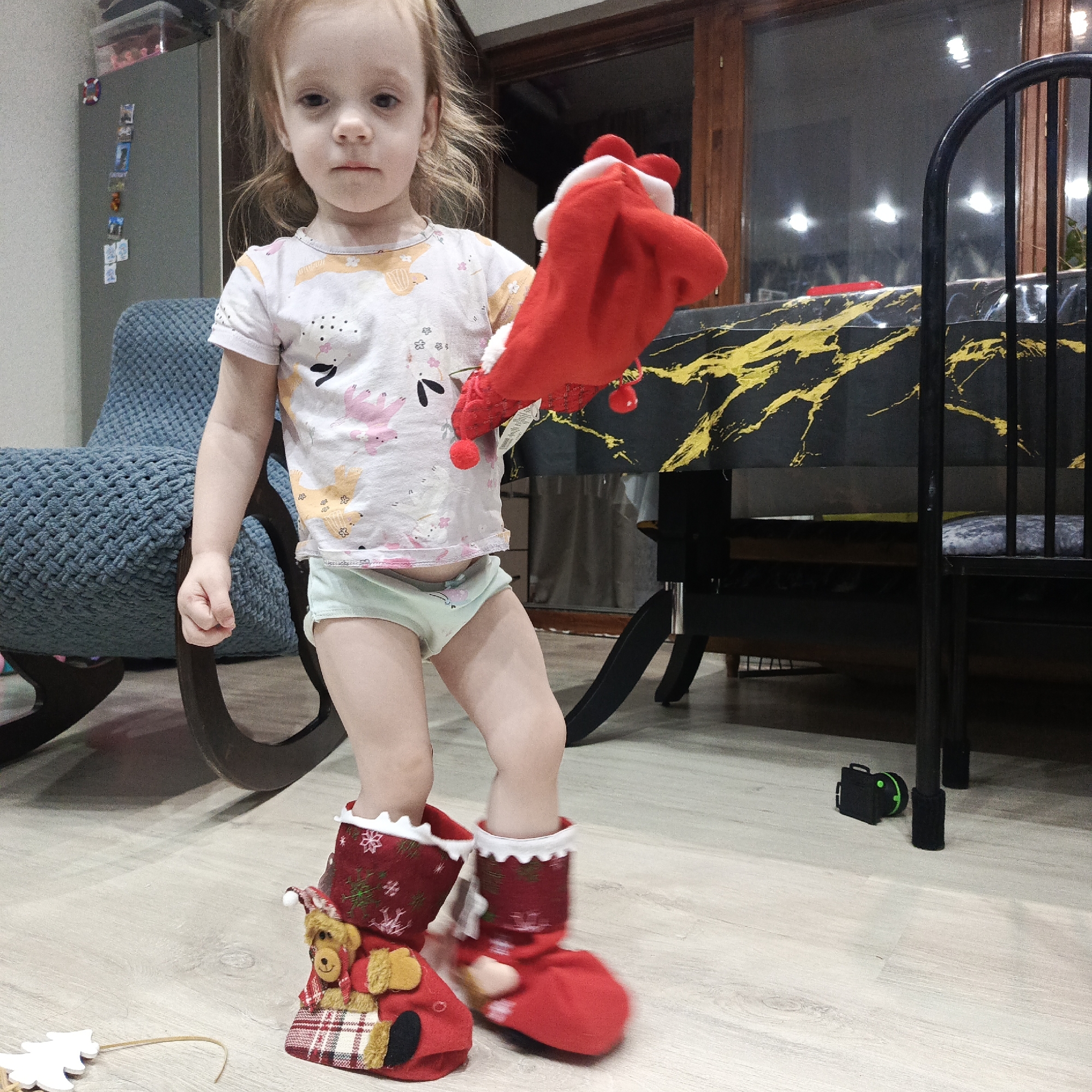 A toddler stands indoors in Марковский, Russia, wearing a patterned t-shirt and underwear, with Christmas stockings on their feet and holding another Christmas stocking in their hand. A blue rocking chair is behind the child on the left, and a table with a black and gold surface is on the right. The floor is light-colored.