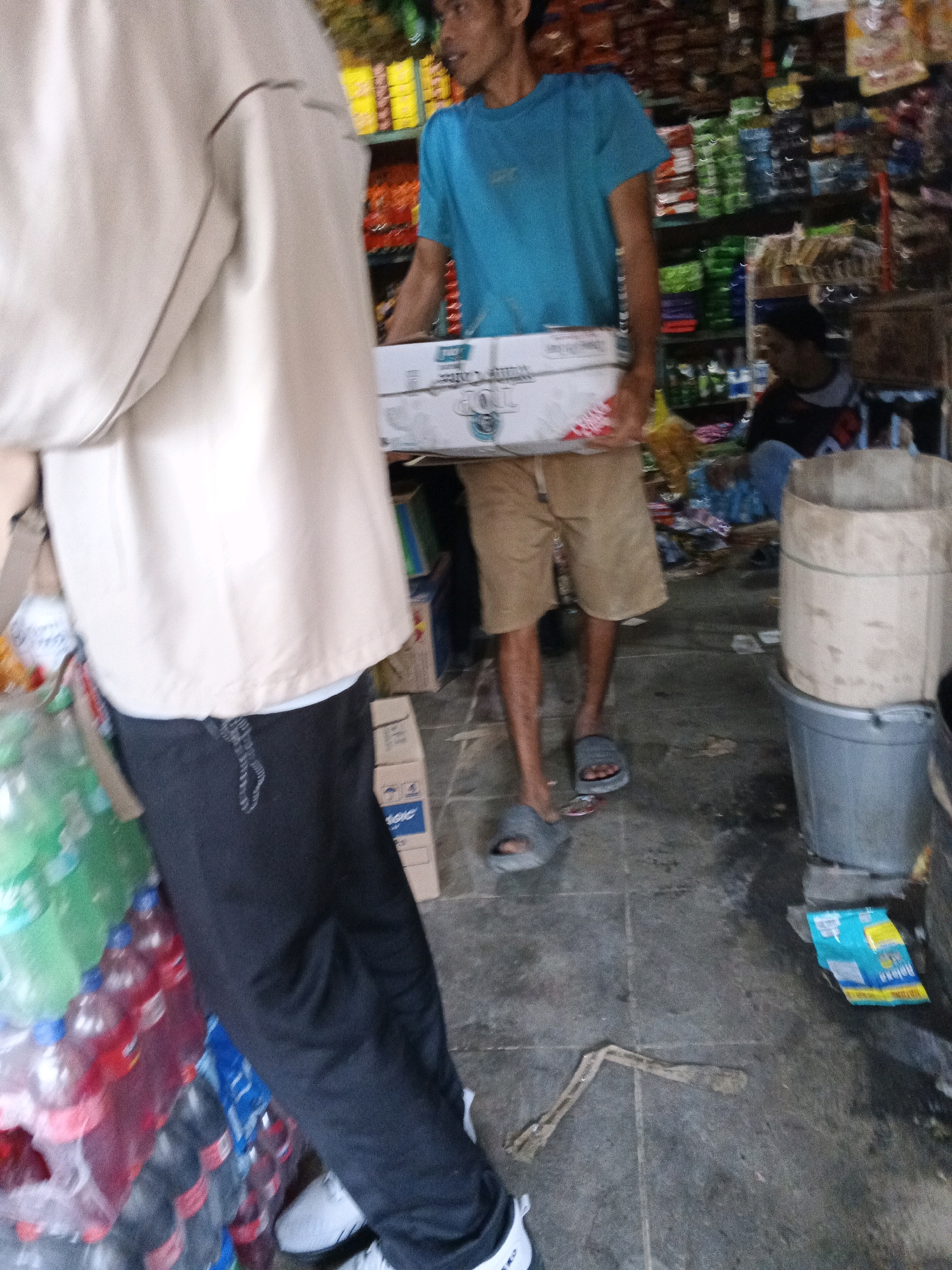 In Warungkiara, Indonesia, a store interior is depicted with shelves stocked with goods. Two individuals are present. In the foreground, a person wearing a beige top and black pants is walking past a stack of bottled drinks. Further back, a second person in a blue t-shirt and khaki shorts is carrying a white box. Another individual, wearing a dark shirt, is seated behind a counter in the background. The scene shows a retail environment with merchandise displayed on shelves, and various items on the floor.