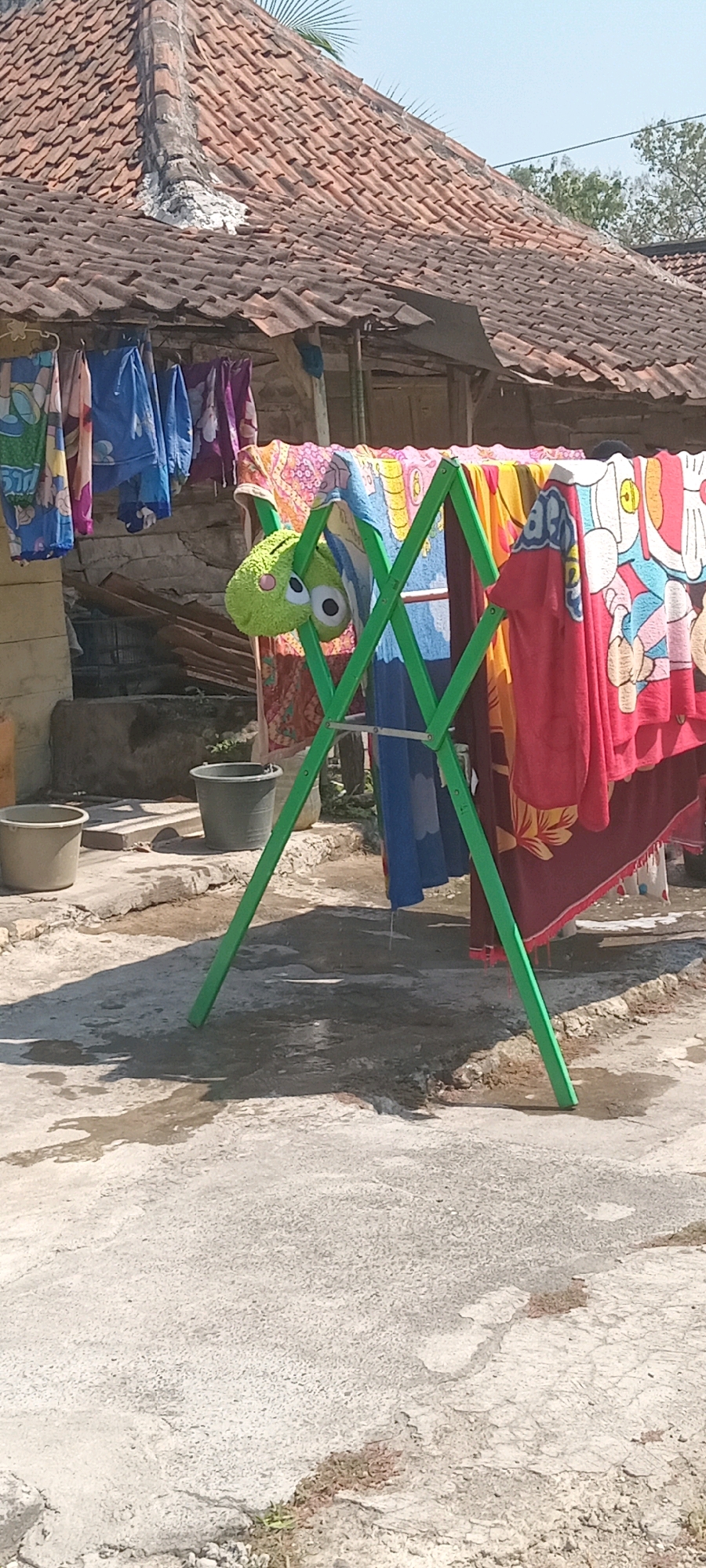 A bright, sunny outdoor scene in Kedukbembem, Indonesia, features a clothesline laden with colorful towels and clothing. A green A-frame clothes rack stands prominently, supporting a variety of items. A small, bright green stuffed toy with large googly eyes is hung from the rack. In the background, a tiled roof of a building is visible, and to the left, two plastic buckets, one light-colored and one dark gray, are placed on the ground. The ground itself is paved with concrete, showing signs of dampness and puddles, indicating recent water exposure, possibly from washing or rain. The overall impression is of a typical domestic laundry area on a clear day.
