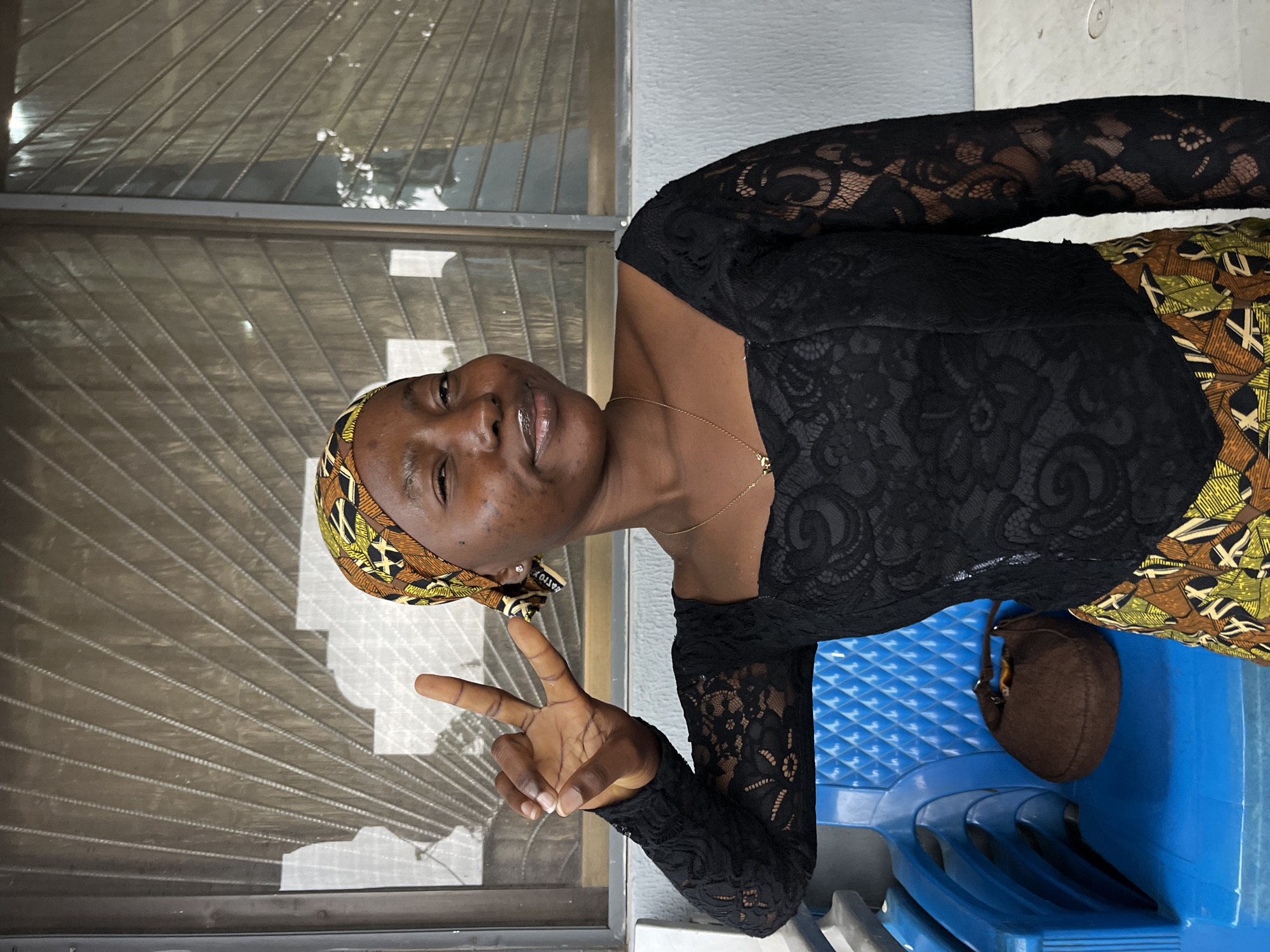 A young Black woman, wearing a black lace top with a colorful headwrap and patterned skirt, poses for a photo. She is winking and holding up two fingers in a peace sign. Behind her, a window with a grate is visible, and to her left, a stack of blue plastic chairs holds a brown handbag. The scene appears to be outdoors, possibly on a porch or balcony.