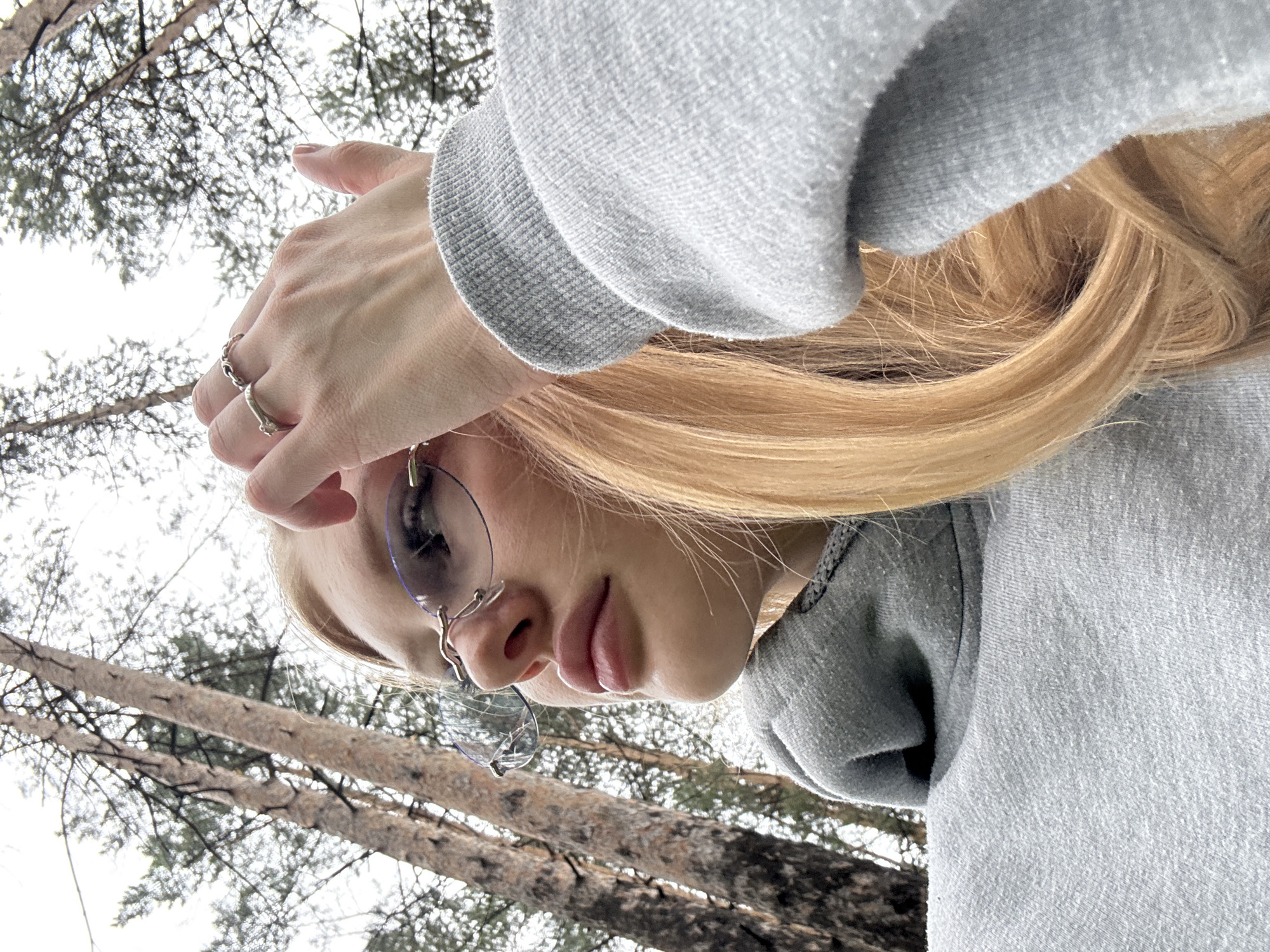 A young woman with blonde hair, wearing a grey hoodie and rimless glasses, is captured in a close-up, low-angle shot. She is outdoors, as indicated by the tall, slender trees and overcast sky in the background, suggestive of a forest or park setting. Her right hand is raised, with fingers touching her forehead, adorned with two silver rings. The image focuses on her face and upper body, with her gaze directed upwards and to the left. The lighting is soft and diffused, consistent with an overcast day. The specific location within the city of Perm, Russia, cannot be determined from the visual information provided.