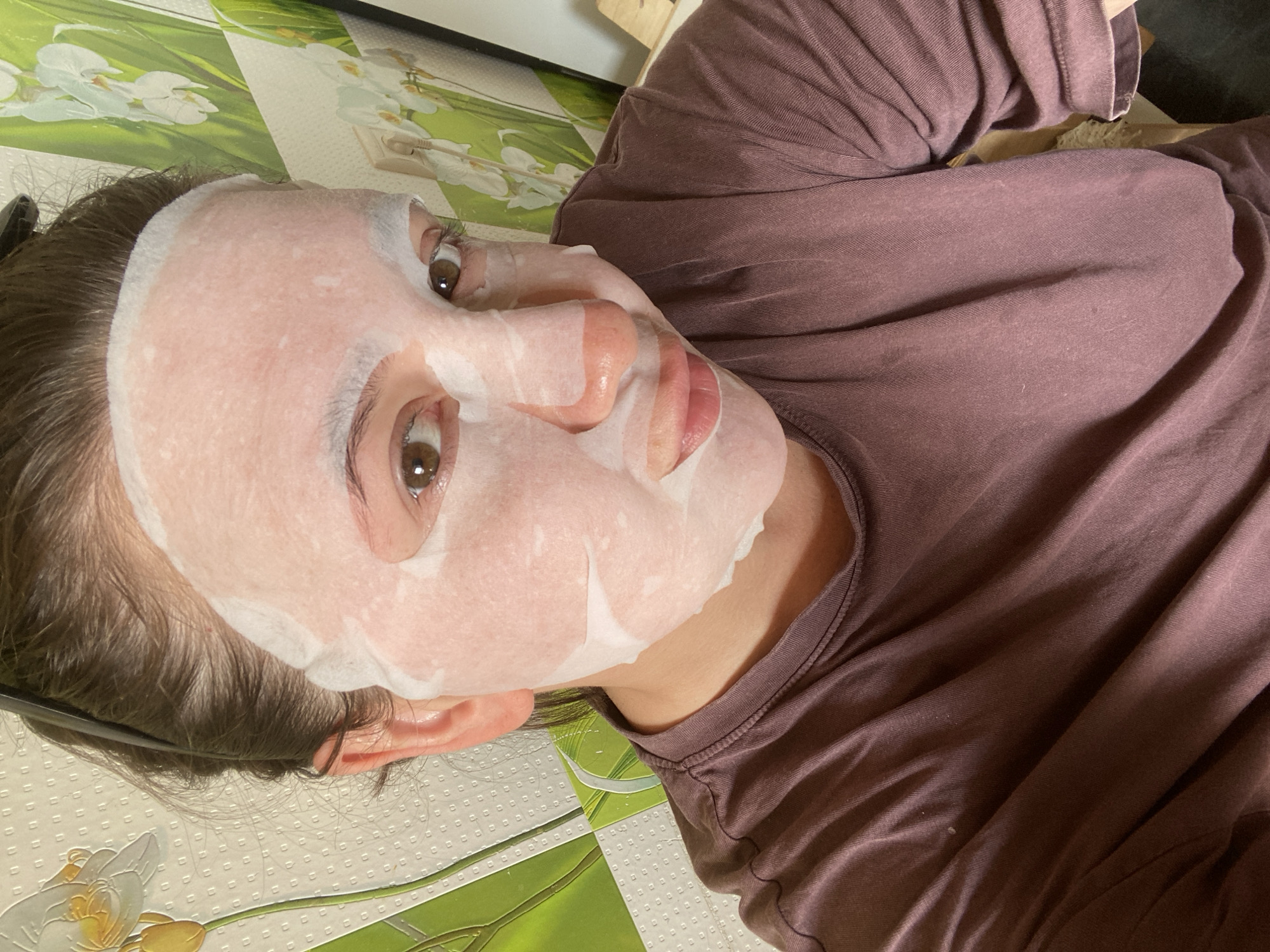 A young woman with brown hair and brown eyes is looking directly at the camera. She is wearing a sheet mask that covers most of her face, with openings for her eyes, nose, and mouth. The mask is white and appears to be made of a thin, absorbent material. The woman is wearing a dark purple t-shirt. The background of the image is a wall with a floral pattern. The overall lighting is bright and even. The image appears to have been taken indoors. This image is not from Chaikovsky, Russia.