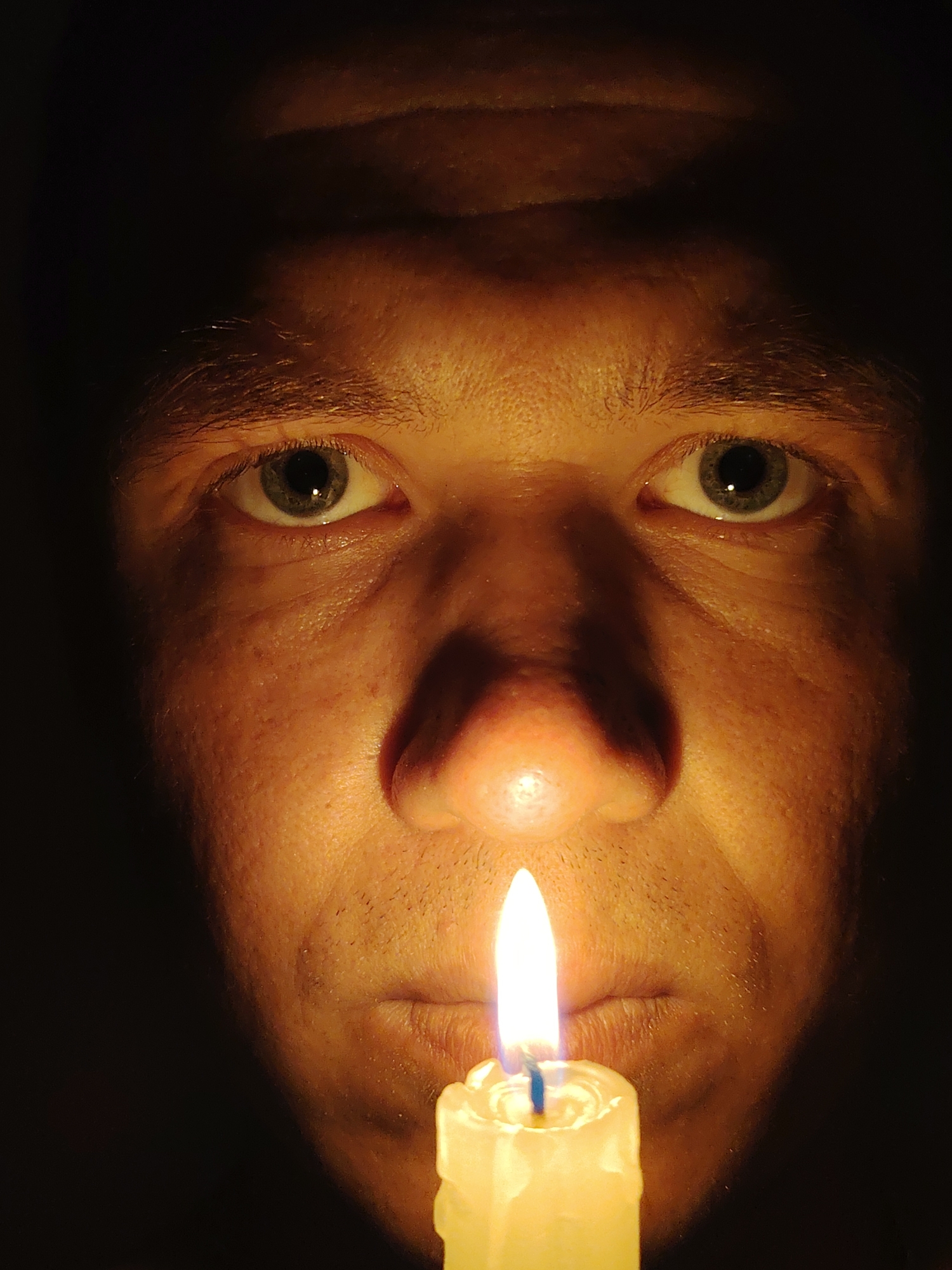 The image is a close-up, low-angle shot of a person's face illuminated by a lit candle held below. The person's eyes are open and looking directly at the camera. The lighting creates strong shadows, particularly on the forehead and sides of the face, emphasizing the contours of the skin. The candle flame is bright white and centered in the lower part of the frame, casting a warm glow upwards onto the person's nose and mouth. The background is entirely dark, suggesting an indoor setting with minimal ambient light. There is no discernible information in the image that would allow for the determination of its location in Chaikovsky, Russia, or any other geographical location.