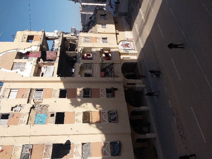A severely damaged building stands beside a wide street with pedestrians. The building's facade shows extensive destruction, with gaping holes where walls and windows used to be. Clothing hangs from some of the remaining structures. The street is partially shaded, and figures walk along its surface. The upper floors of the building reveal significant structural damage, exposing the interiors.