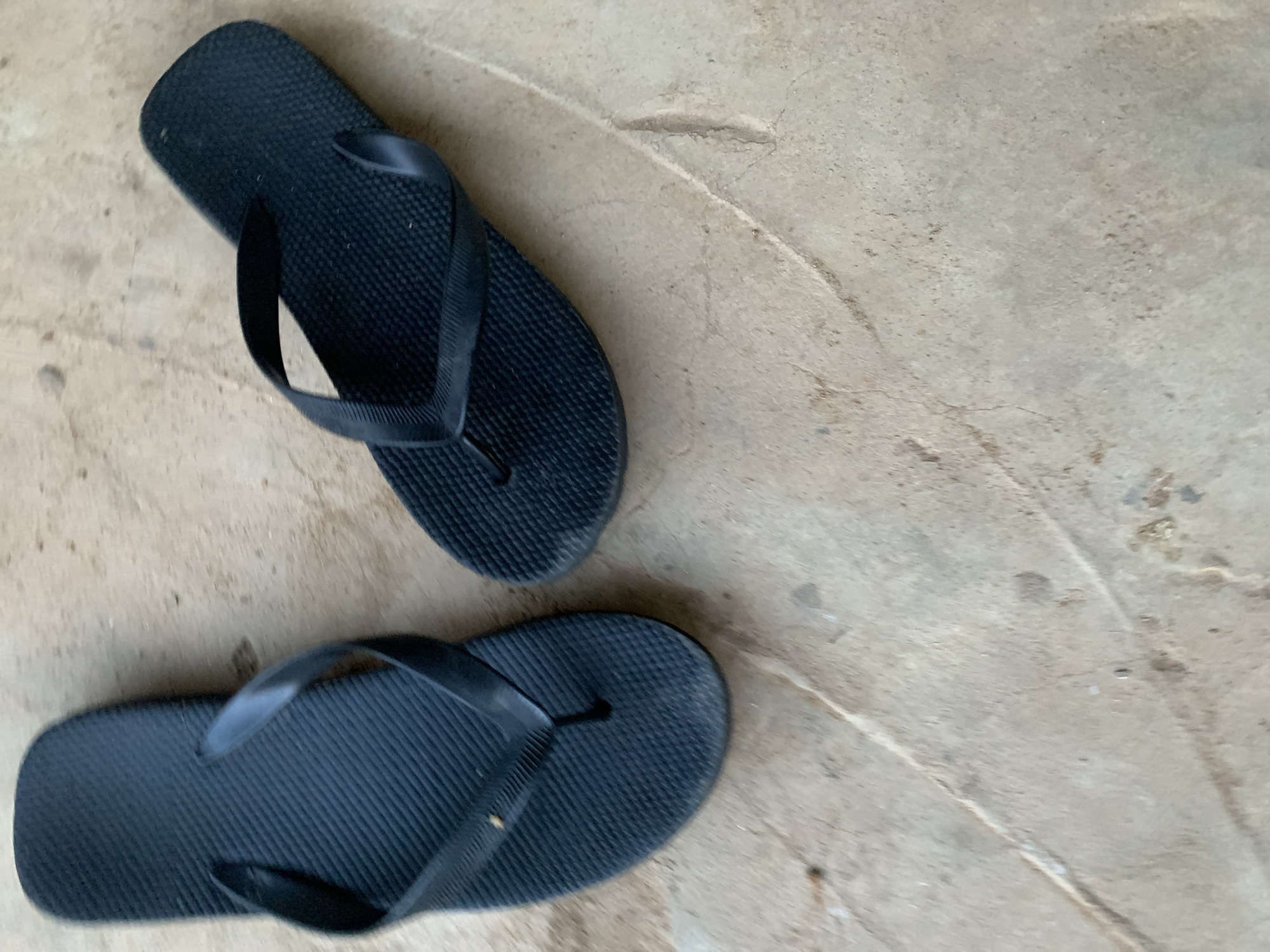 Two black flip-flops (thong sandals) are laid casually on a light beige or off-white ground surface. One flip-flop is positioned in the upper-left portion of the frame, oriented diagonally upwards, while the other rests towards the bottom-center, pointing diagonally to the right. Both flip-flops are solid black, featuring textured soles with small, raised ridges or bumps, and smooth, matte straps.
The ground appears to be a concrete or hard-packed earth surface, showing signs of age and wear. It is characterized by a rough texture, minor cracks, subtle discolorations, and patches of dust or dirt. Faint, curved indentations are visible across the ground, suggesting irregularities in its surface. The lighting is soft and diffused, indicating a daytime setting without harsh shadows, possibly under an overcast sky or in indirect light. There is no human activity depicted, nor any visible text in the image. The scene conveys a simple, casual outdoor environment, consistent with a setting in Keffi, Nigeria.