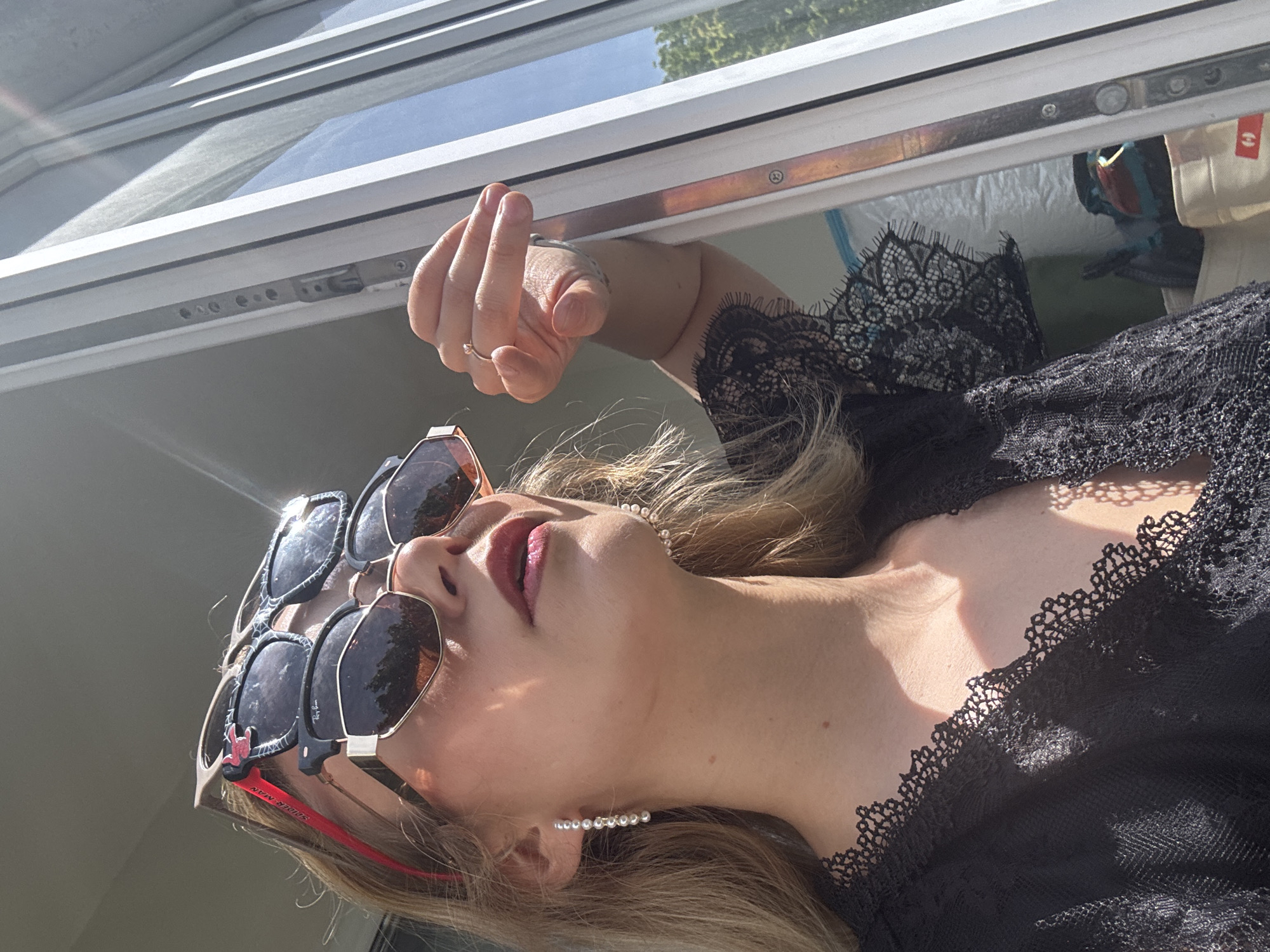 A woman is seen in a low-angle, portrait-oriented close-up. She is wearing two pairs of sunglasses stacked on her nose. The top pair has black frames with a spider-man sticker and the bottom pair has a gold and red frame. The woman's lips are a dark red. She is also wearing a pearl necklace and pearl earrings. She is wearing a black lace top or dress. Her left hand is visible, with a ring on one of the fingers. The background is a window frame, with a partially open window and foliage visible. No evidence suggests this photo was taken in Riga, Latvia.