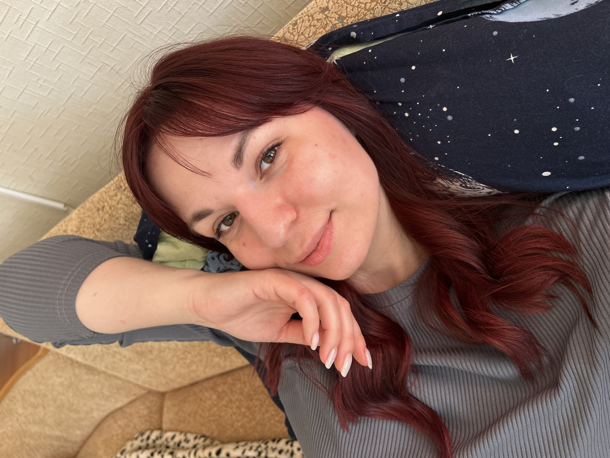 A woman with auburn hair is reclining on a beige sofa. Her hair is styled in loose waves. She is wearing a gray, ribbed long-sleeved shirt. Her right hand is resting on her cheek. Her nails are long, oval-shaped, and painted with a clear iridescent polish. A dark blue pillow with white star-like patterns is behind her head. There is also a lighter blue patterned fabric near her head. Part of a brown surface is visible at the top left of the frame. There is a leopard print material in the bottom left corner. The picture is taken at a close-up angle.
There is no indication the image was taken in Фоки, Russia.