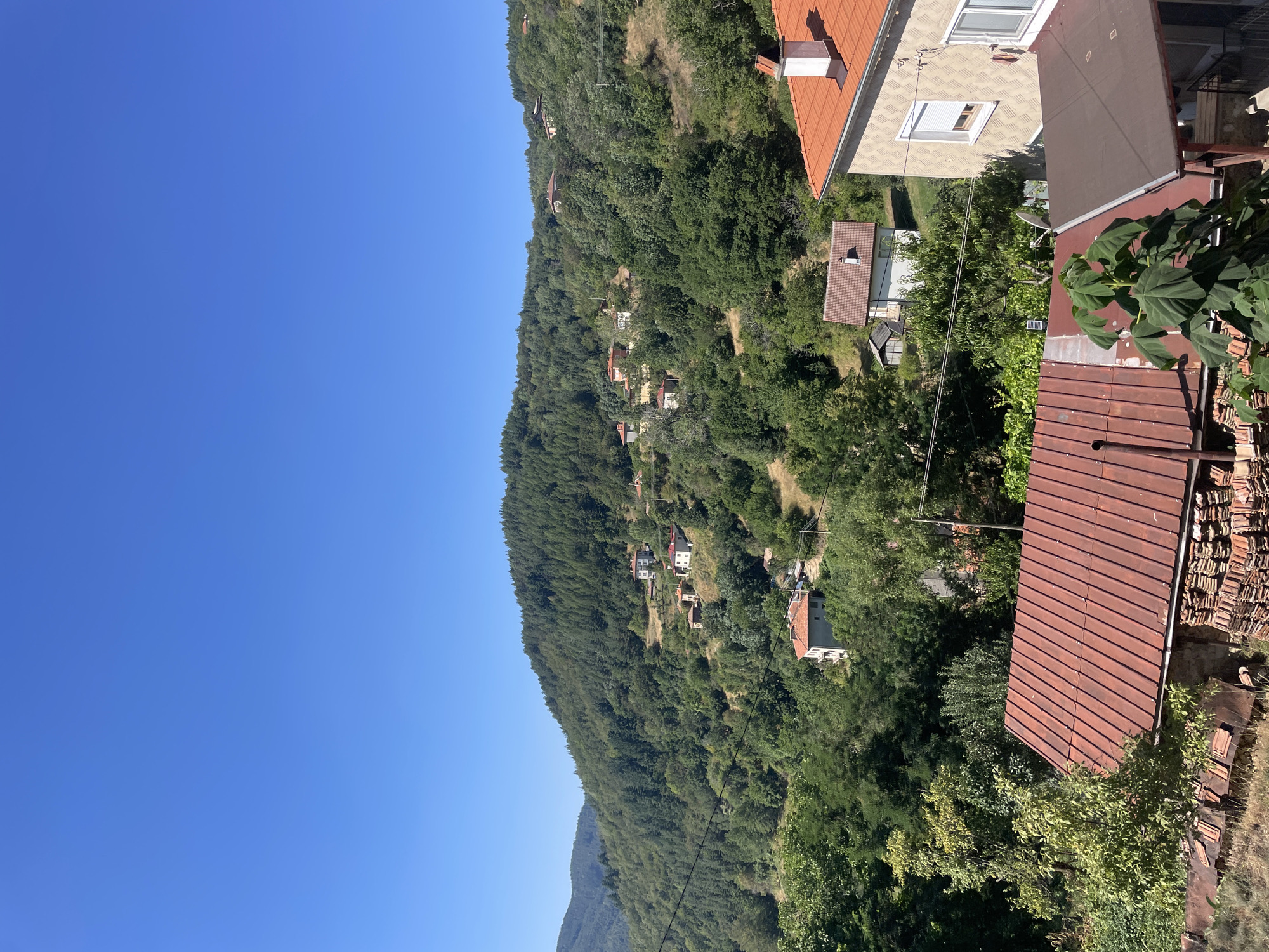 The image depicts a residential area nestled within a lush, hilly landscape. The foreground features several rooftops, predominantly covered in weathered, reddish-brown tiles. To the left, a pile of stacked roof tiles is visible. In the middle ground, a cluster of houses with red-tiled roofs are scattered across the slopes, surrounded by dense green trees. The houses vary in architectural style, with some appearing to be older stone structures and others more modern constructions. Further in the background, the hills are densely covered in coniferous and deciduous trees, creating a natural, verdant backdrop. The sky is a clear, bright blue, indicating a sunny day. The overall scene conveys a sense of rural tranquility in the village of Davidkovo, Bulgaria.