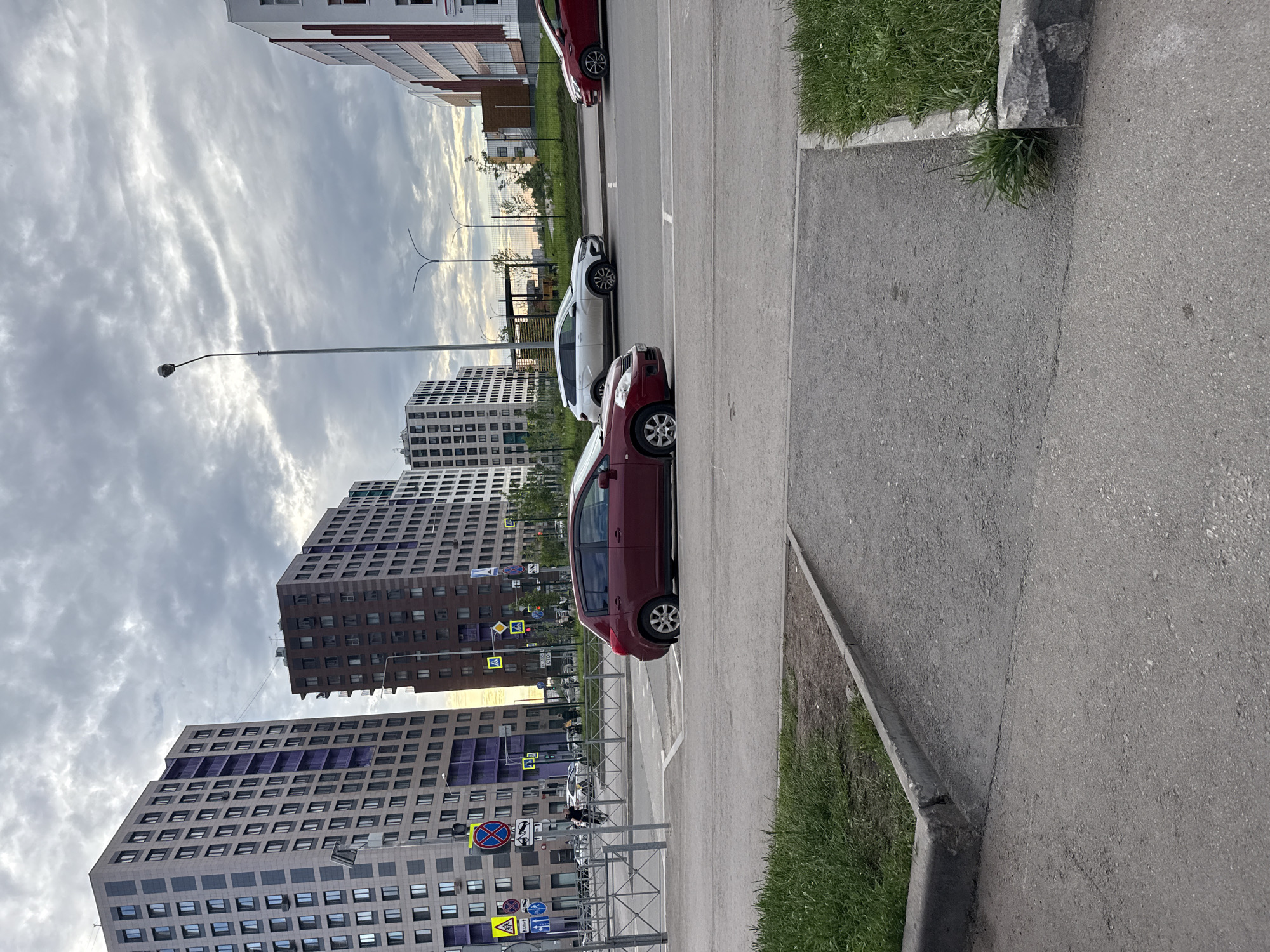 The image shows a street scene in Perm, Russia, with several tall apartment buildings lining a roadway. The sky is overcast with dramatic, cloudy formations, suggesting a potentially stormy or late afternoon/early morning time of day. Several cars are parked along the side of the road, including a red hatchback and a white car. A street lamp is visible in the foreground, and metal fencing separates the sidewalk from a grassy area. Various road signs are also present, including traffic signs and parking restrictions. The architecture of the buildings is modern, with many windows and balconies. One building has purple accents around some of its windows. The overall mood is somewhat somber due to the cloudy sky, but the presence of parked cars and buildings suggests an active urban environment. No people are clearly visible in the image.